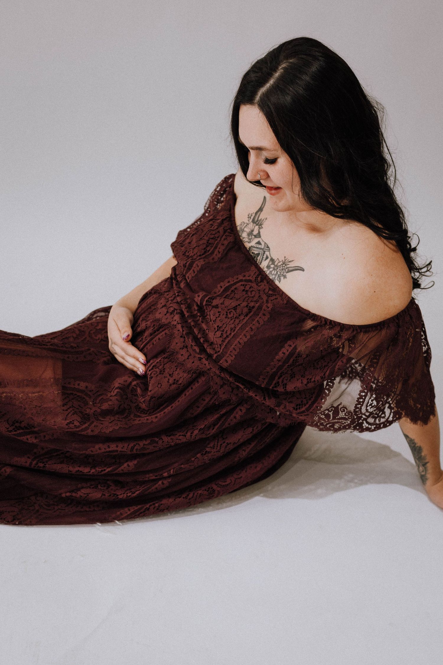 A figure in a burgundy lace dress poses gracefully on a light background while looking down at the flowing fabric.