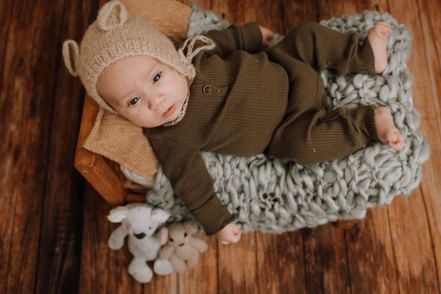Baby in brown outfit and bear bonnet lying on chunky knit blanket with stuffed toy.