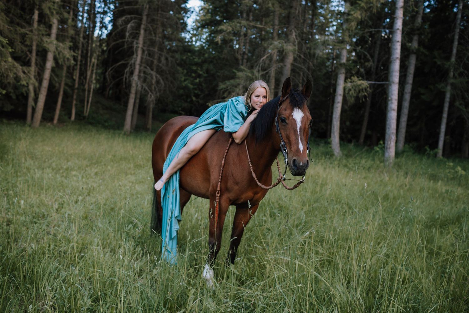 Rider in flowing turquoise dress draped over brown horse in wooded meadow.