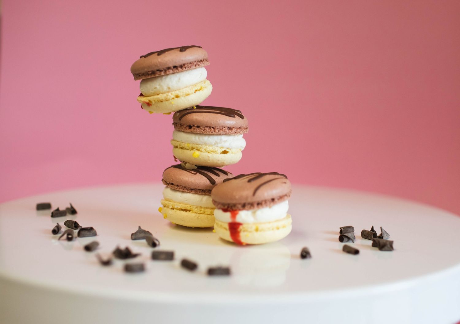 Stacked chocolate and vanilla macarons with chocolate chips scattered on white surface against pink background.