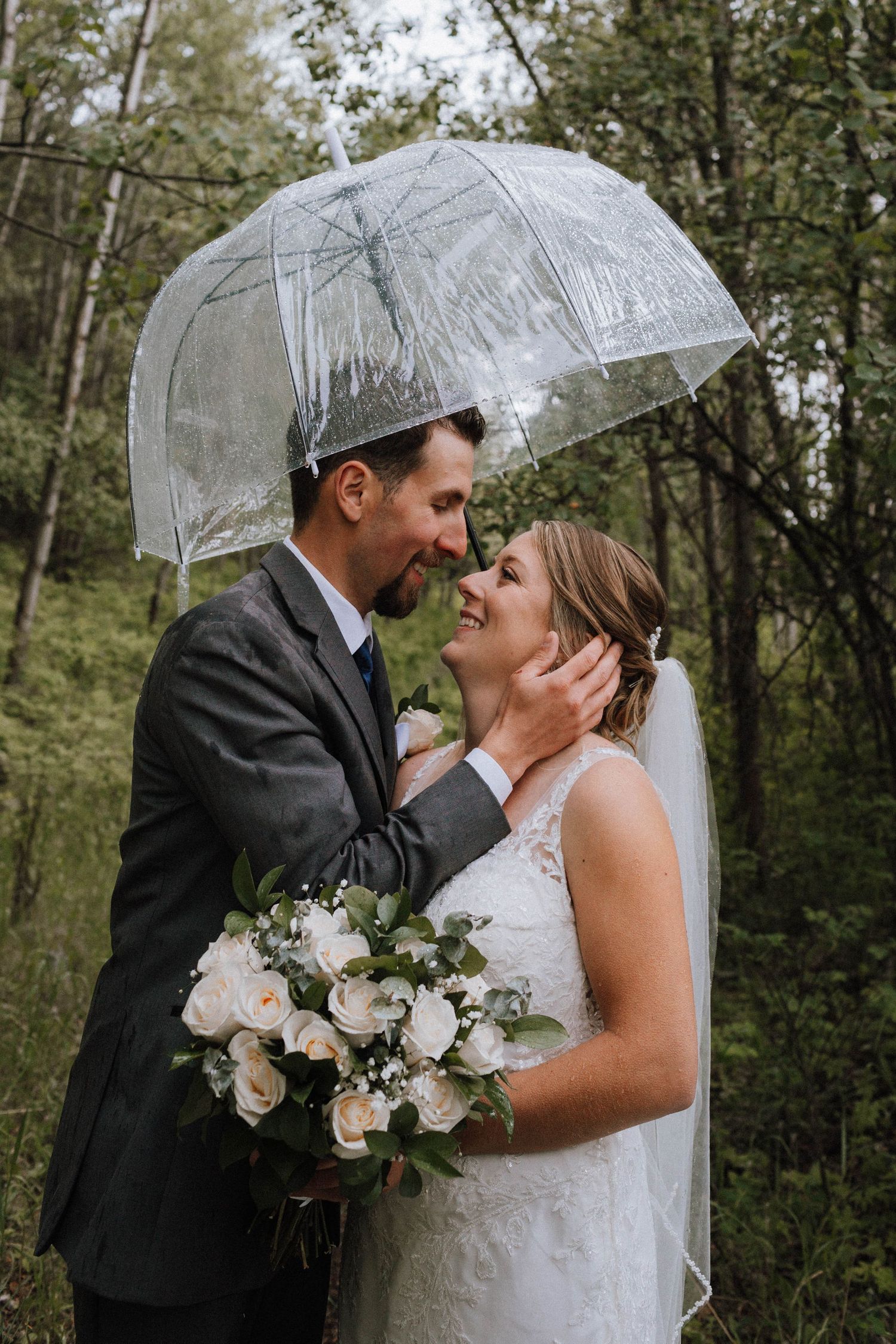 Romantic wedding couple sharing an intimate moment under a clear umbrella in a forest setting with white bouquet.