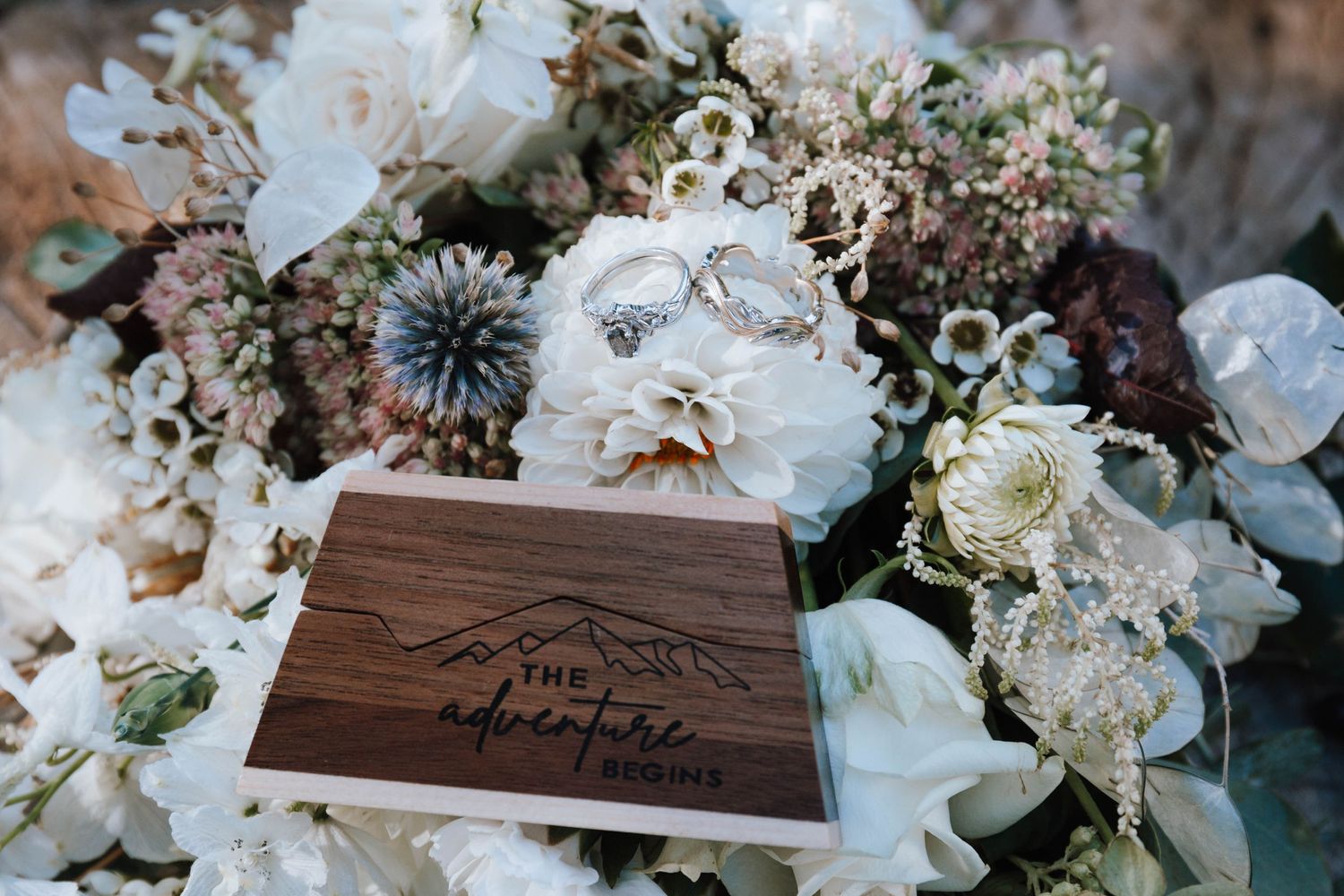 Wooden wedding ring box surrounded by delicate white and purple floral arrangements.