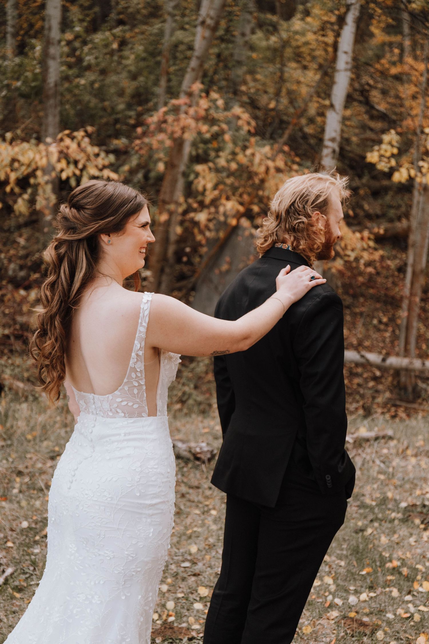 A bride in a lace wedding dress with her hand on her partner's shoulder in an autumn forest.