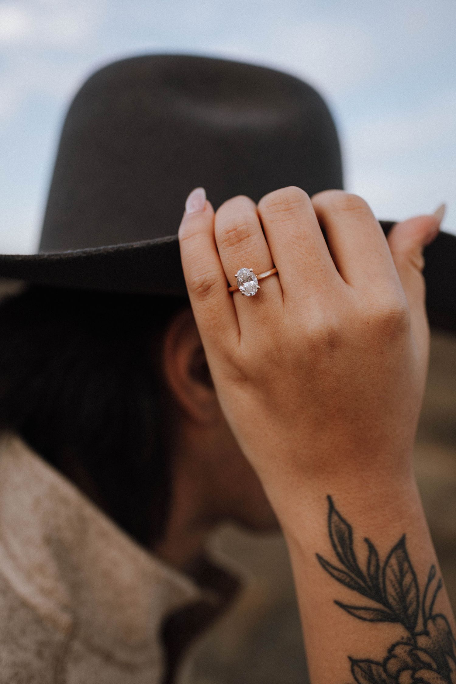 A hand wearing a solitaire diamond engagement ring with a floral tattoo on the wrist.