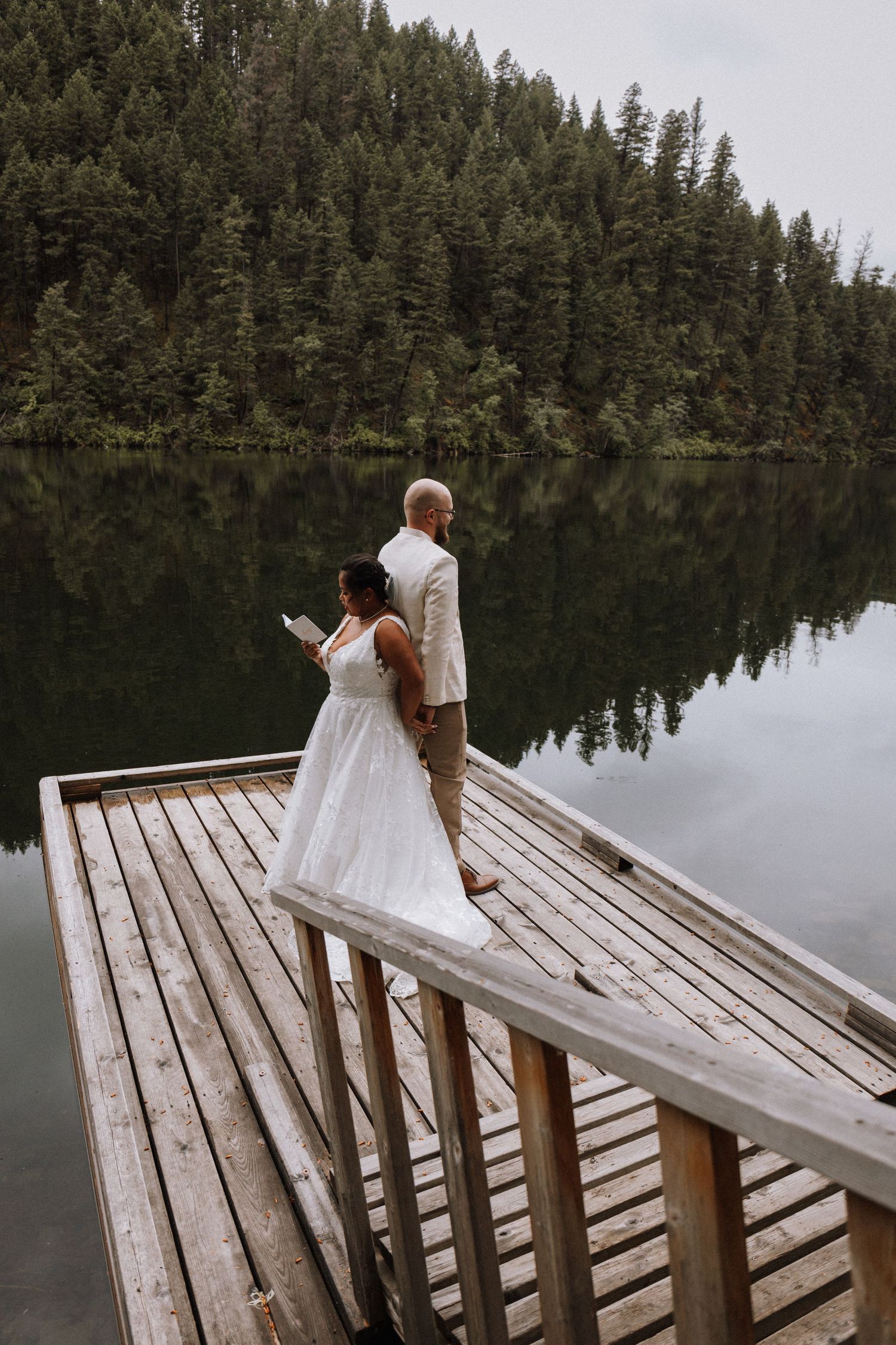 Bride and groom stand on a wooden dock overlooking a serene forest lake during their intimate outdoor elopement ceremony.