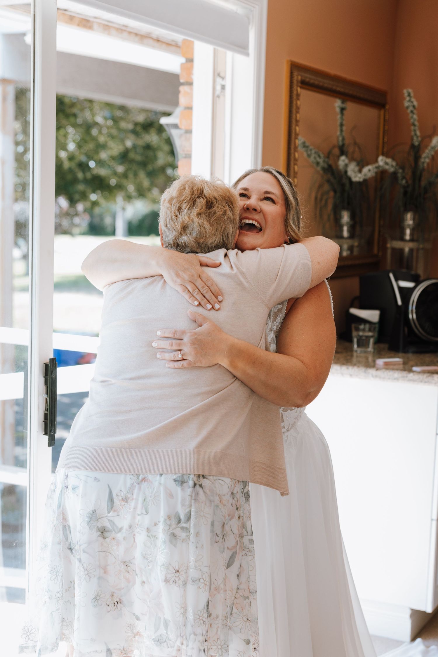 A joyful bride in a floral gown shares a warm embrace with an older woman in a bright indoor setting.