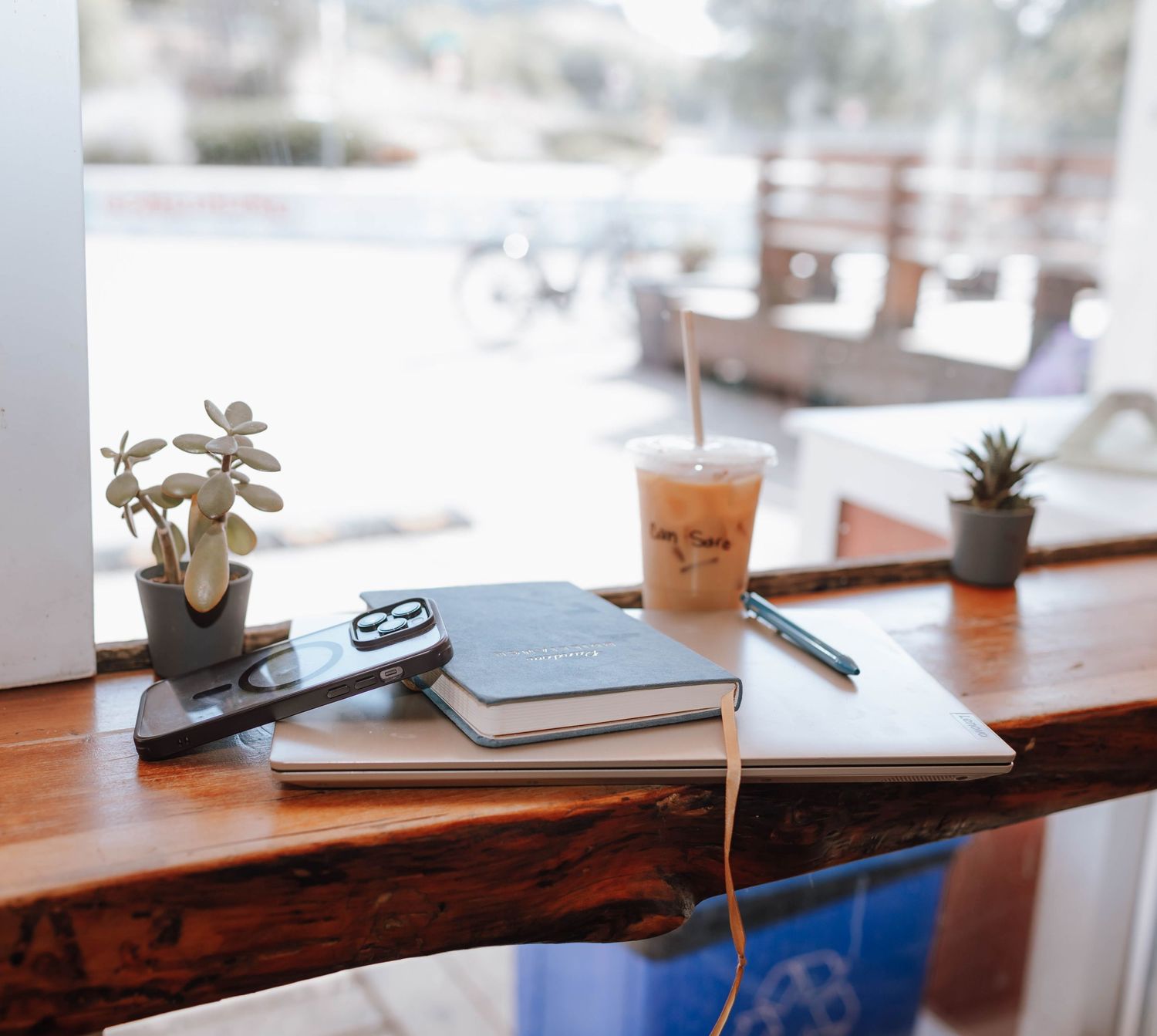 Workspace setup at cafe with iced coffee, notebook, and small potted plants.