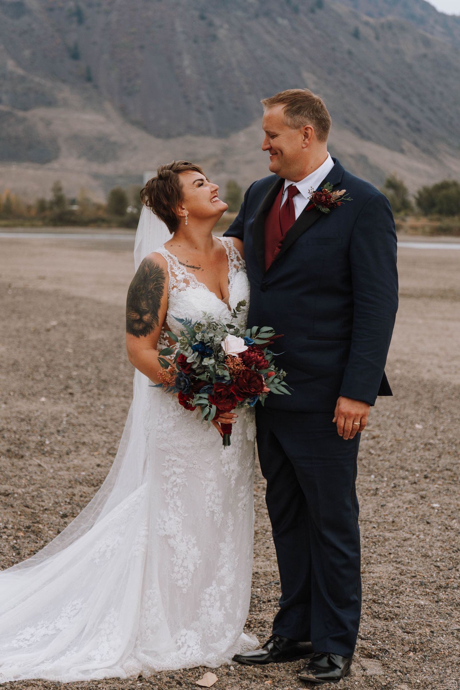 A couple in wedding attire embraces while standing on desert terrain with mountains in the background.