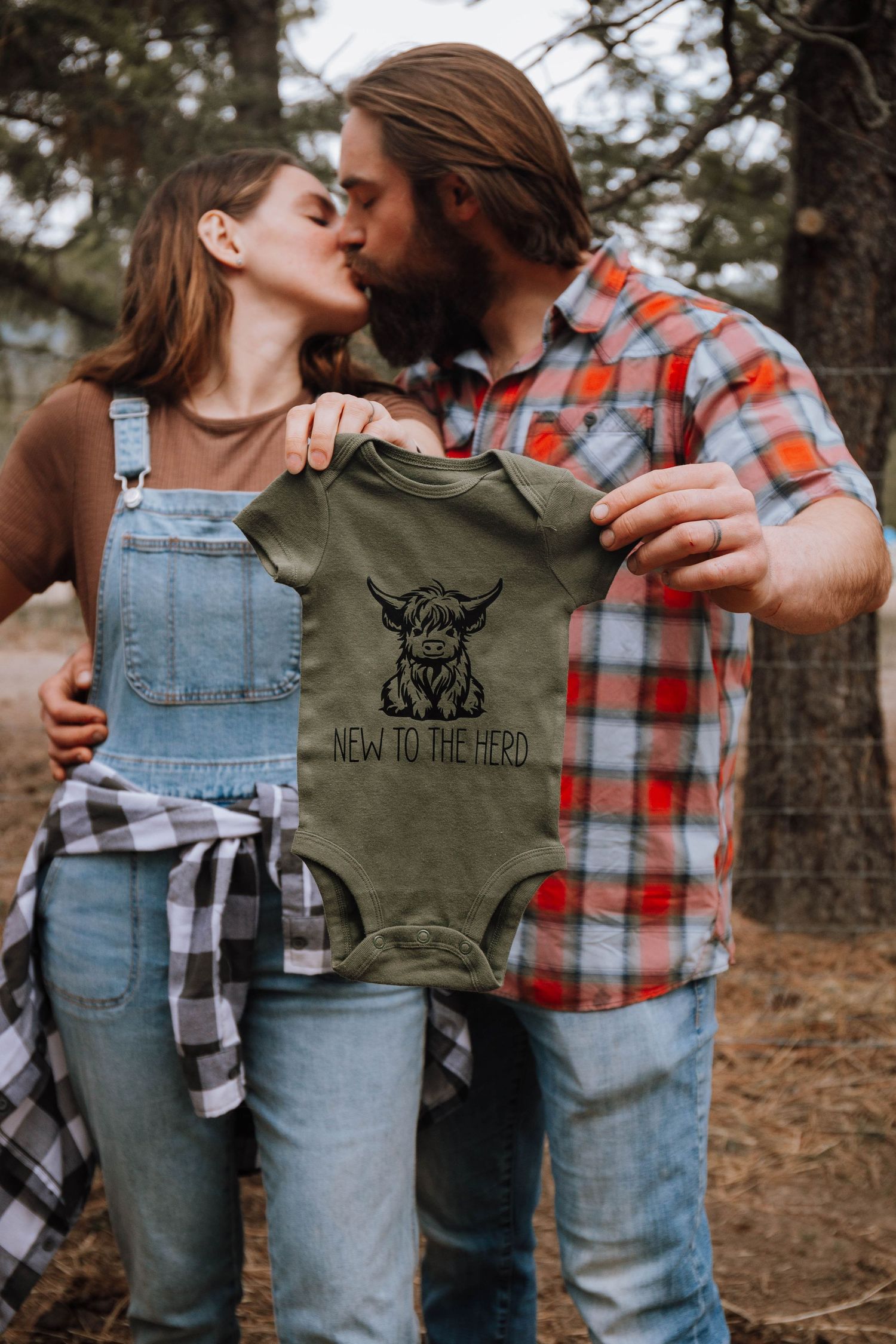 A couple outdoors holding up a khaki green baby onesie with an animal design.