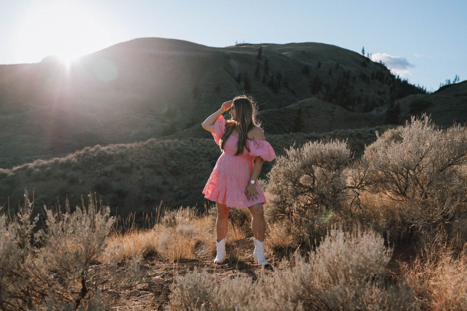 A person in a pink dress poses against a sunlit mountainous backdrop with sagebrush.