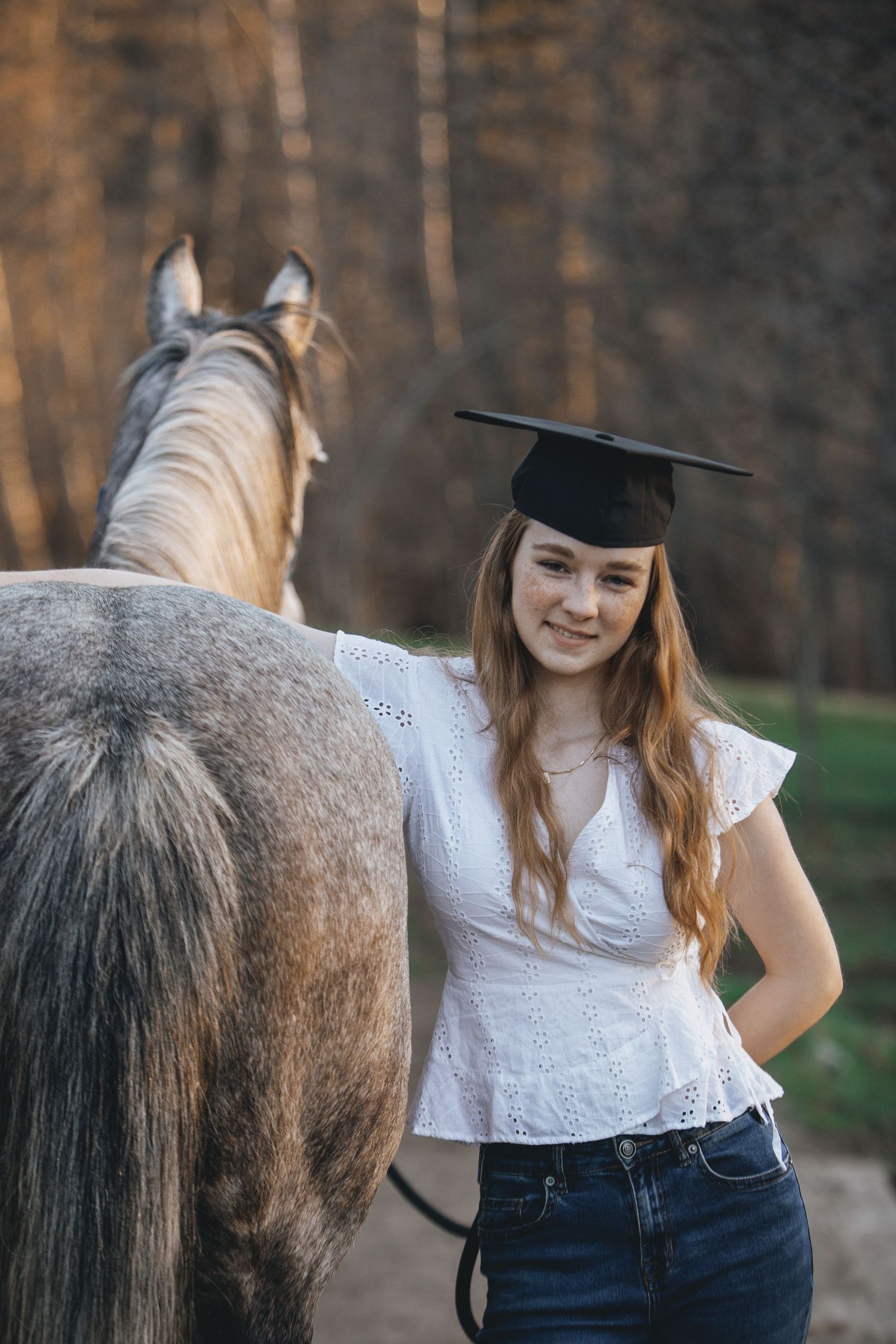 Graduate in cap and white top stands beside gray horse in forest setting.