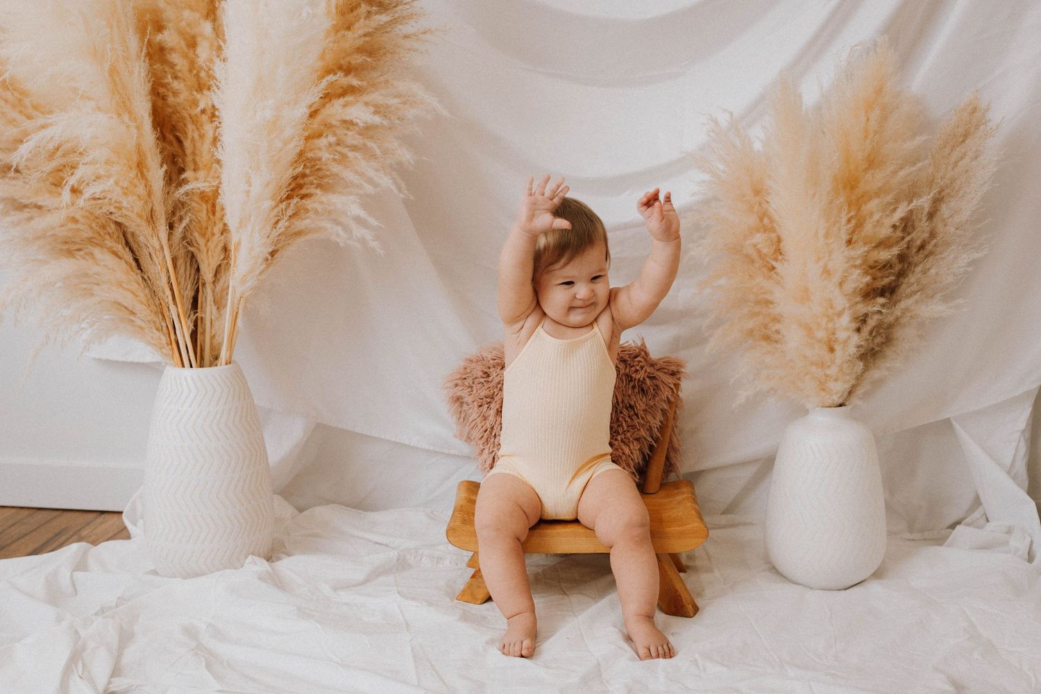 Happy baby in cream romper with raised arms sitting among pampas grass arrangements.