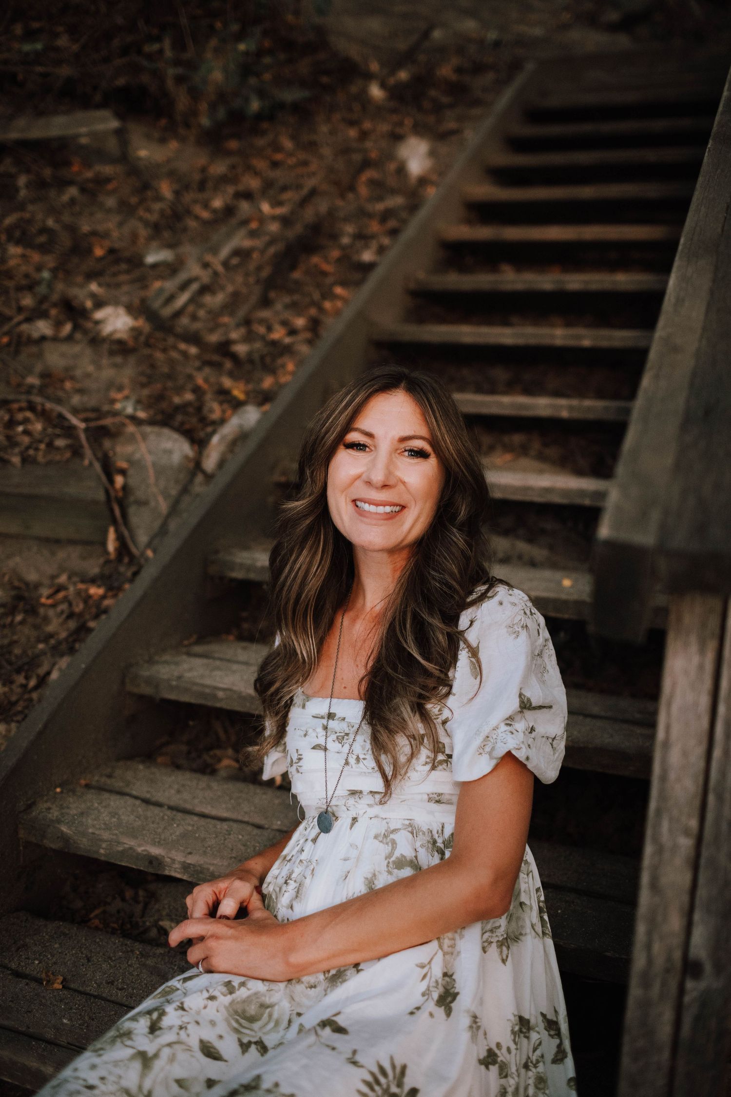 Person in floral dress sitting on rustic wooden stairs surrounded by autumn foliage.