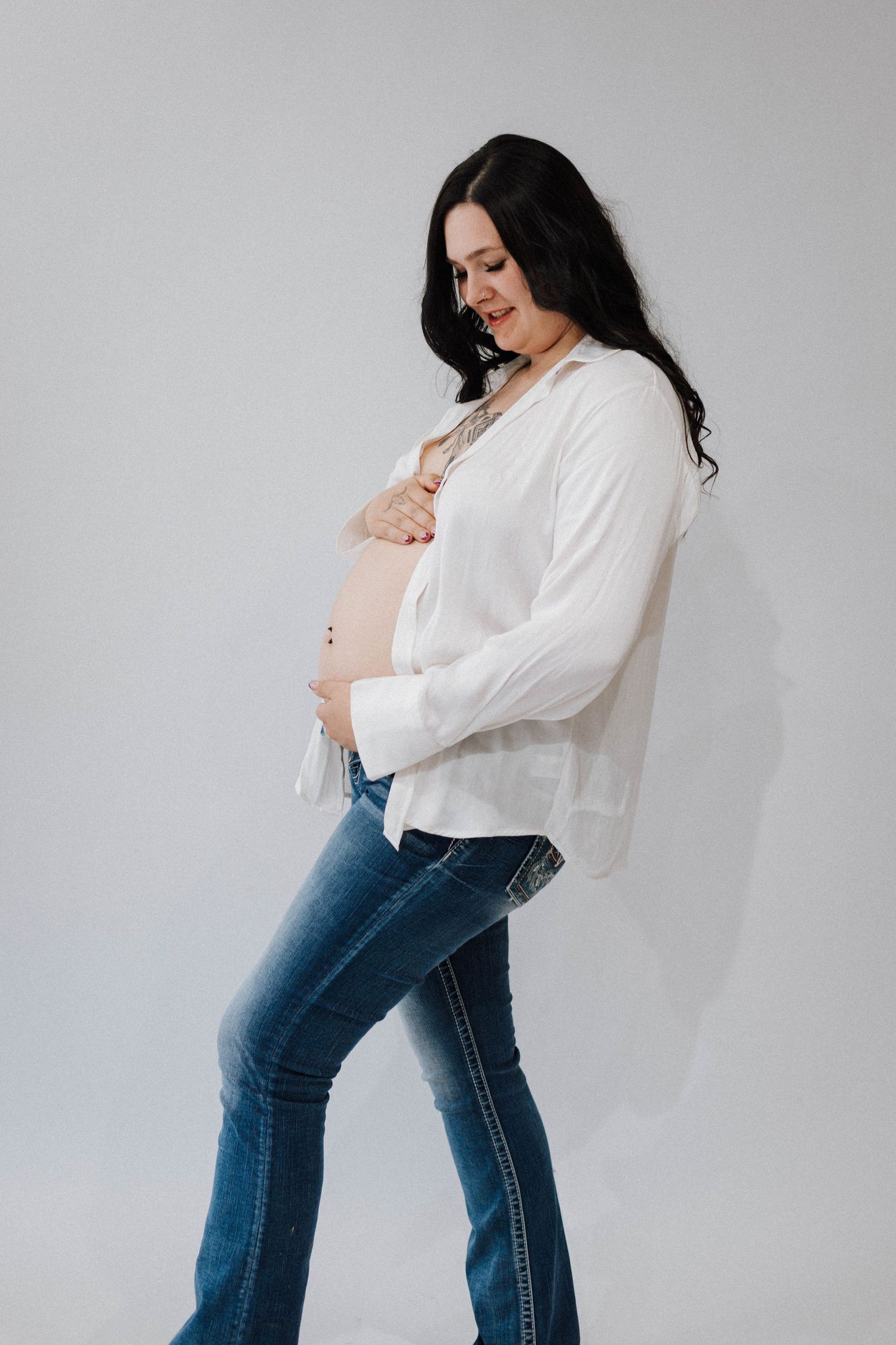 Expectant woman in white blouse and jeans poses against gray background in casual maternity photoshoot.