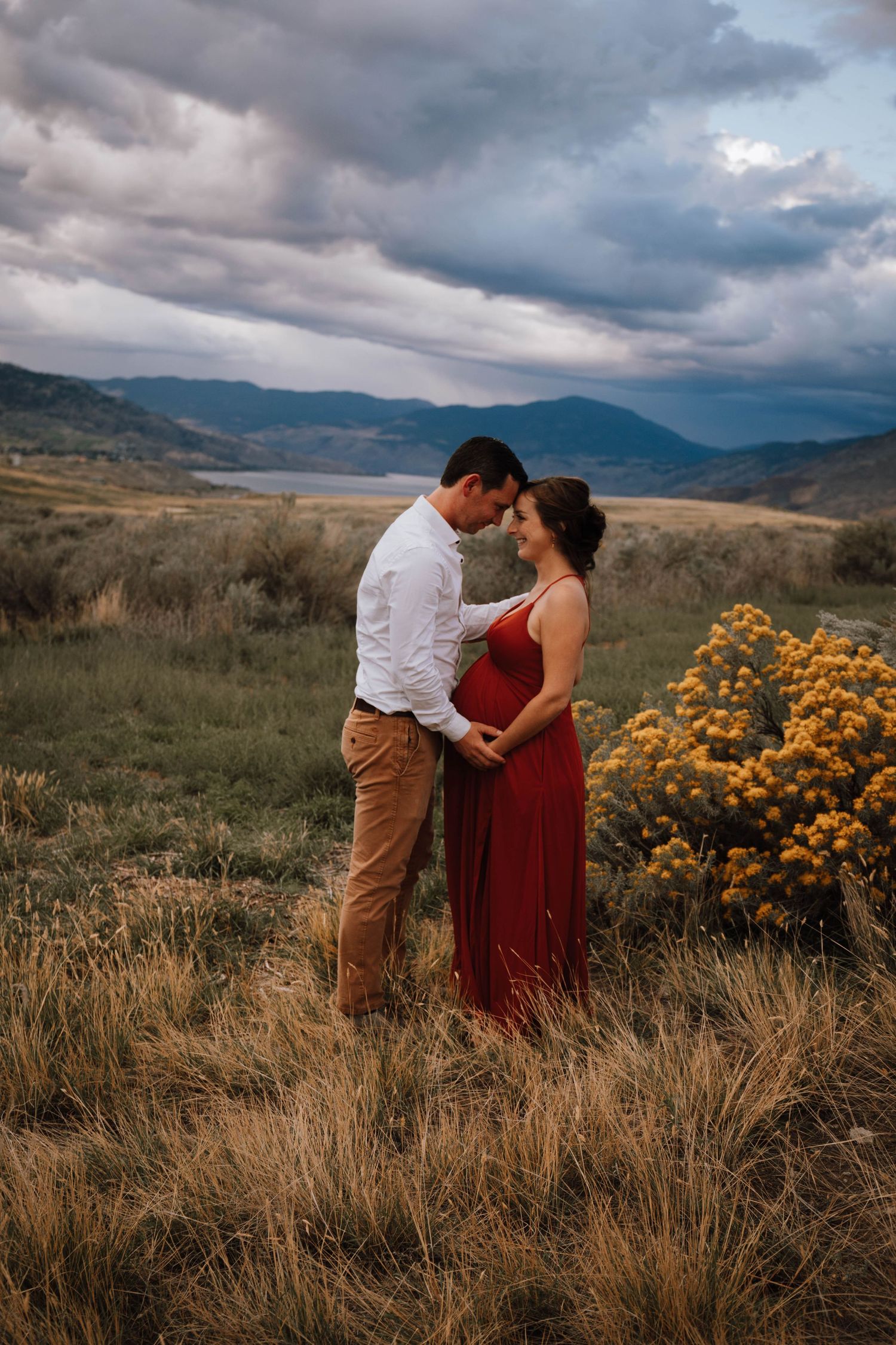 A couple shares an intimate moment in a mountainous field with dramatic stormy clouds and yellow wildflowers at sunset.