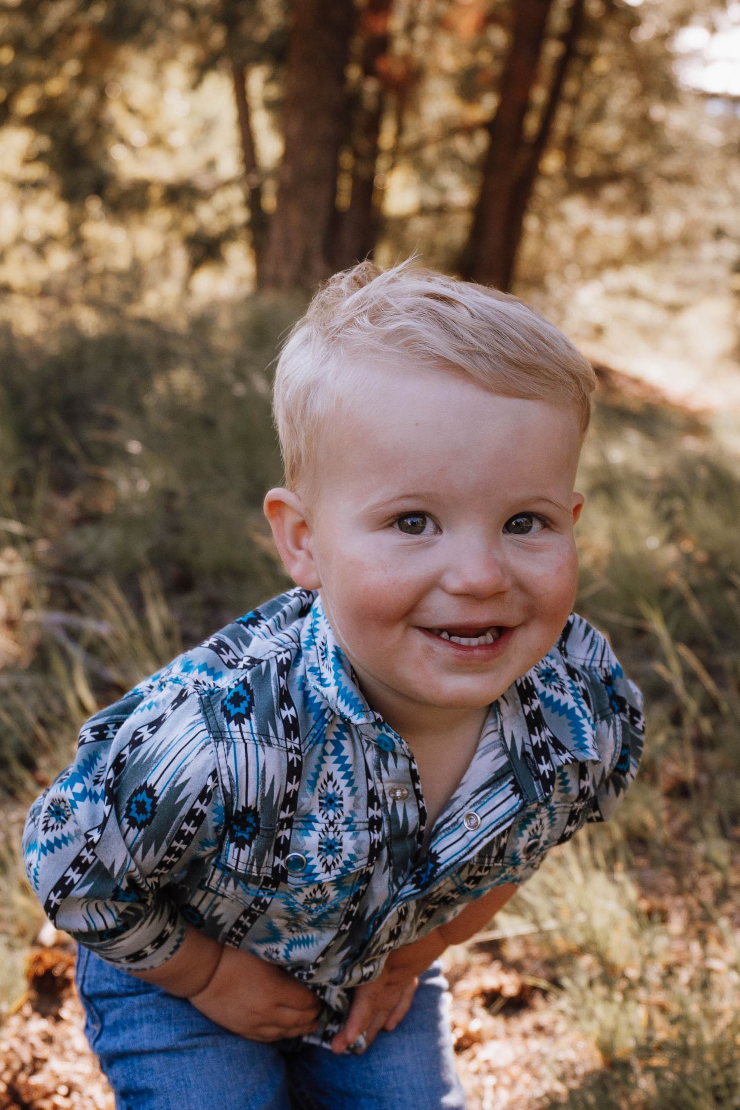 A young child in a blue patterned Hawaiian shirt bends forward with a bright smile amid a wooded outdoor setting.
