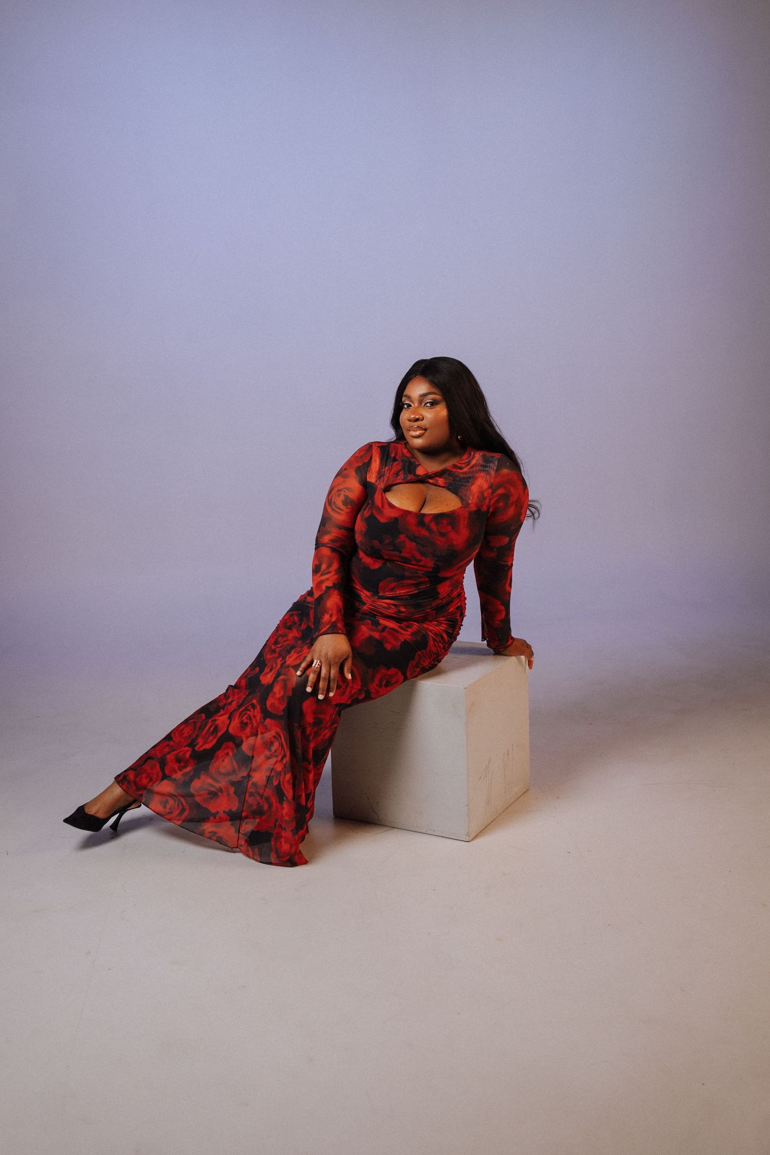 Woman in red floral long-sleeve maxi dress with cutout detail sits on white cube in studio photo.