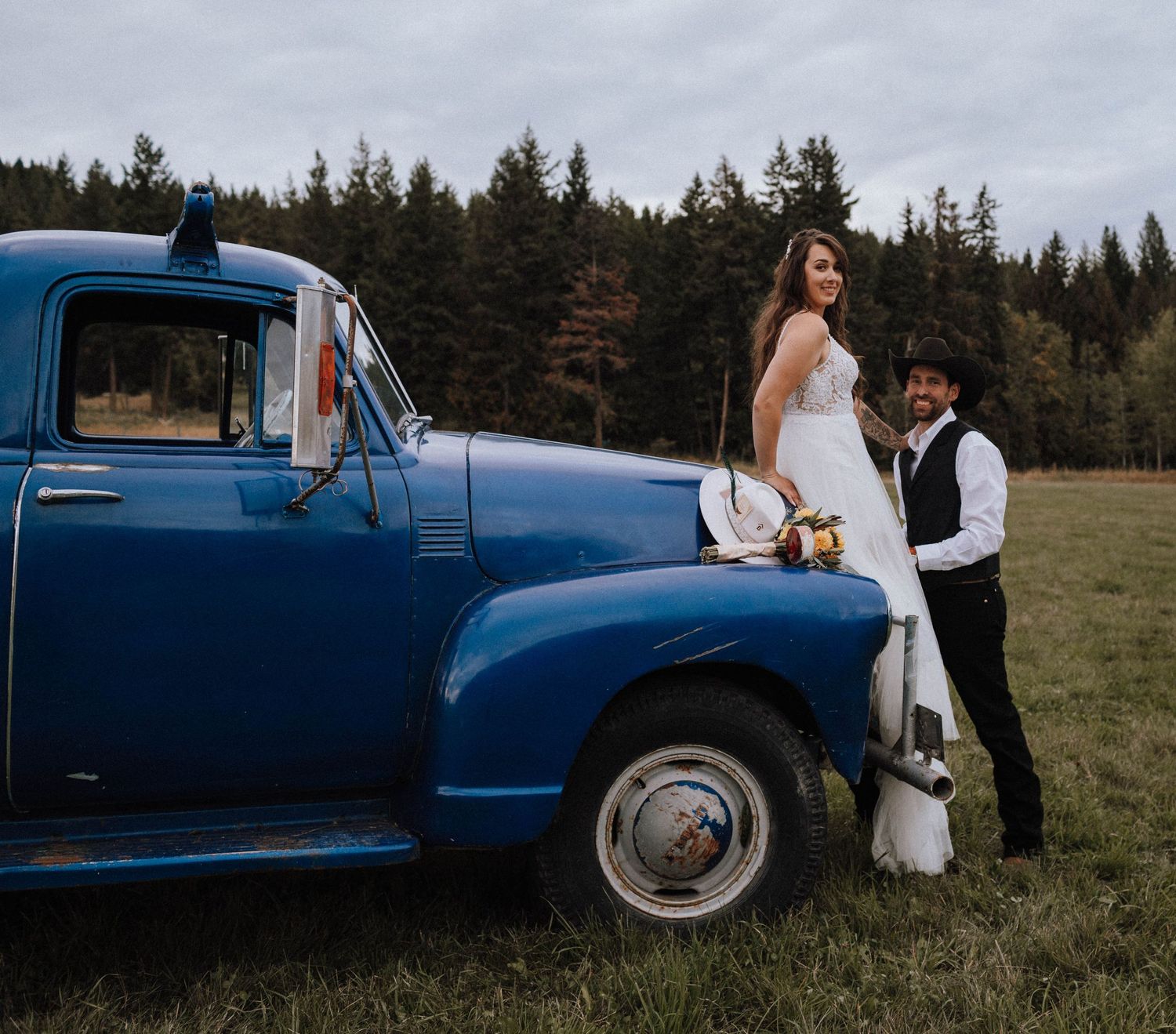 A couple poses with a vintage blue pickup truck in a rustic outdoor setting.