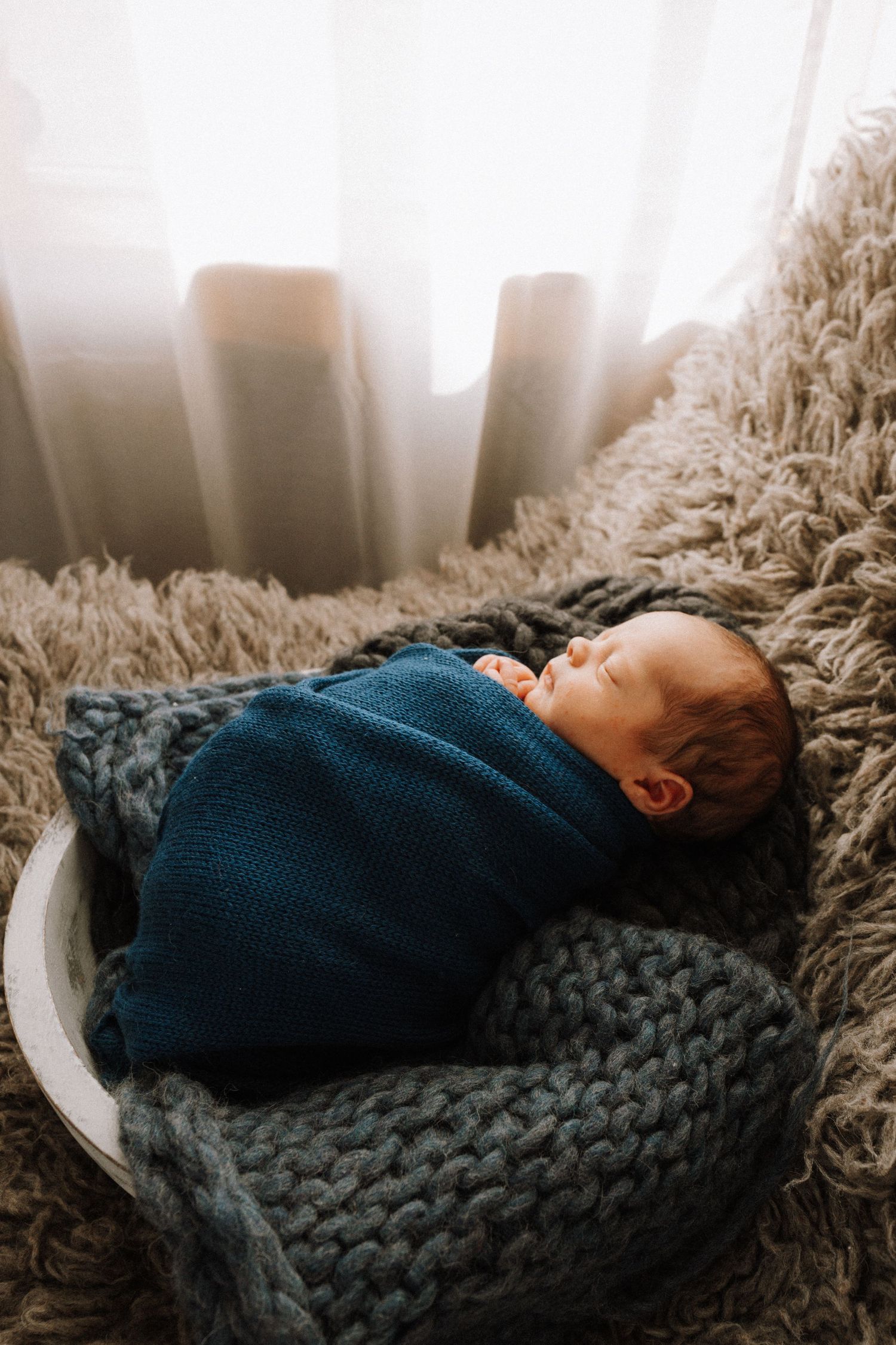 A sleeping newborn wrapped in a navy blue sweater rests on a chunky knit blanket and plush rug near a sunlit window.