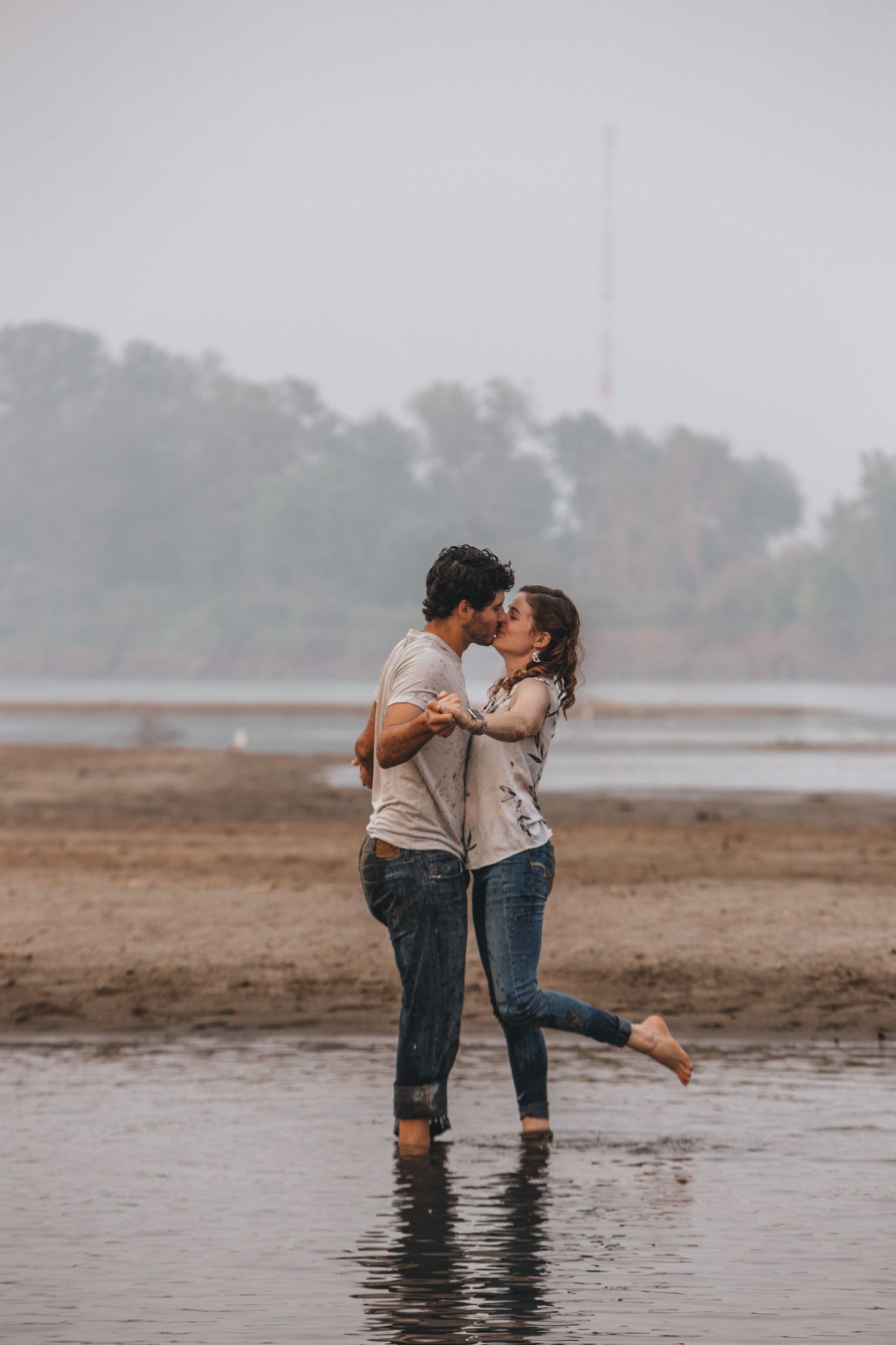 A romantic couple shares playful moments while standing in shallow water during an overcast day.