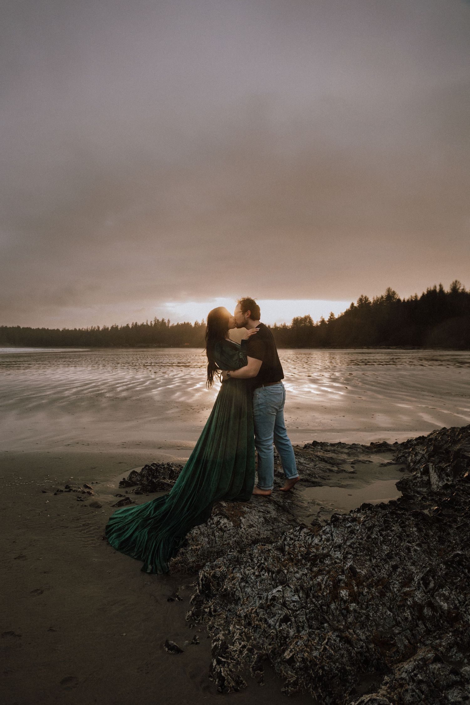 Couple embracing on a rocky beach at sunset, woman in flowing green gown, golden light reflecting on calm water.