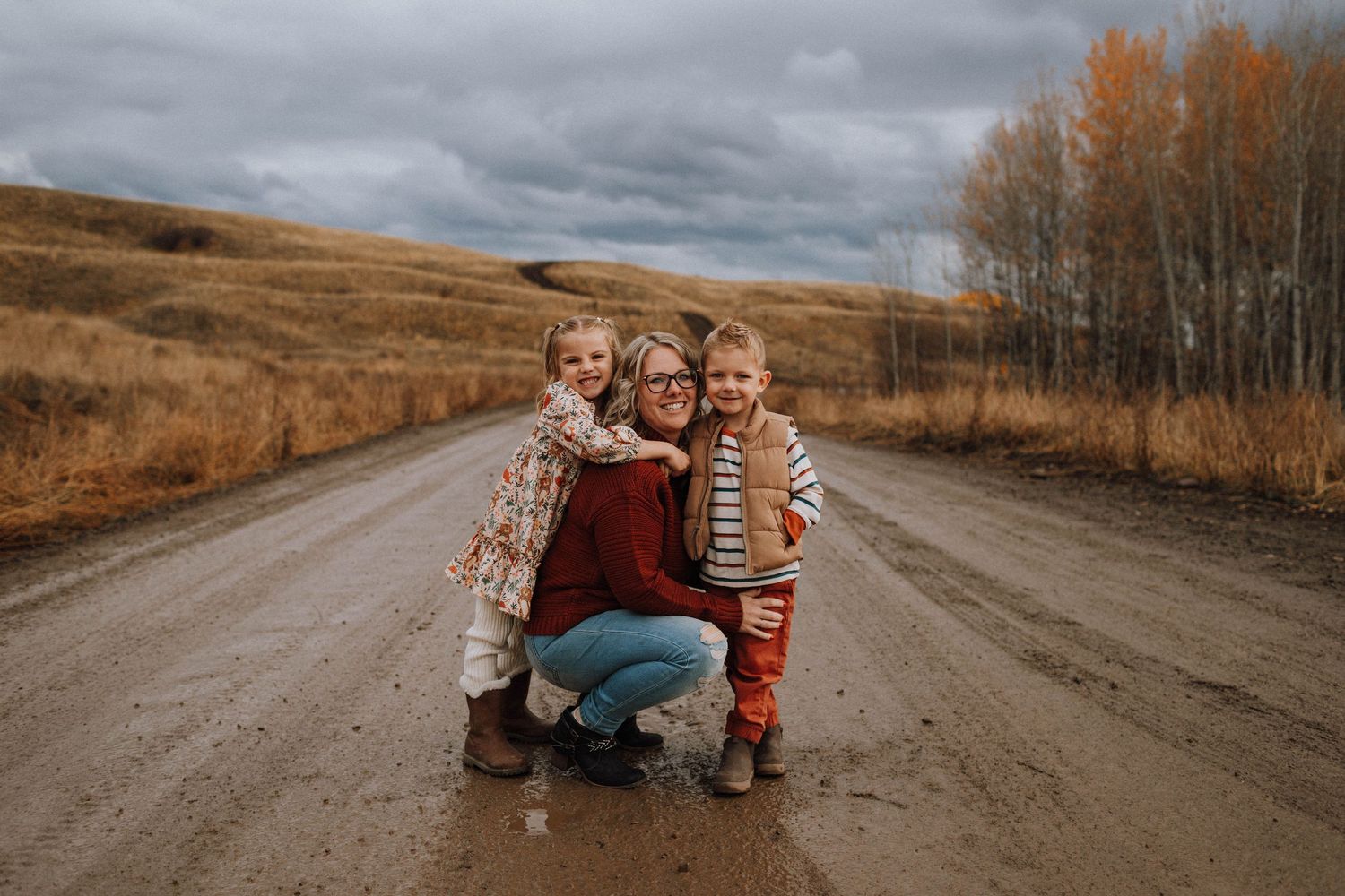 A mother crouches on a rural dirt road hugging her two young children amid autumn trees and golden fields.