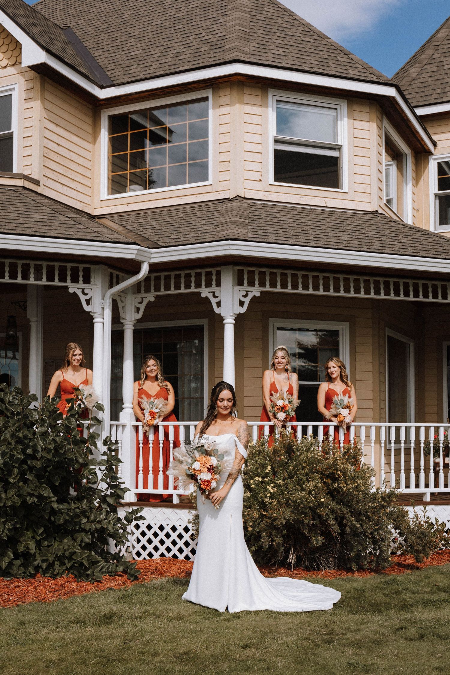 A wedding party poses in front of a Victorian-style house with a wraparound porch on a sunny day.