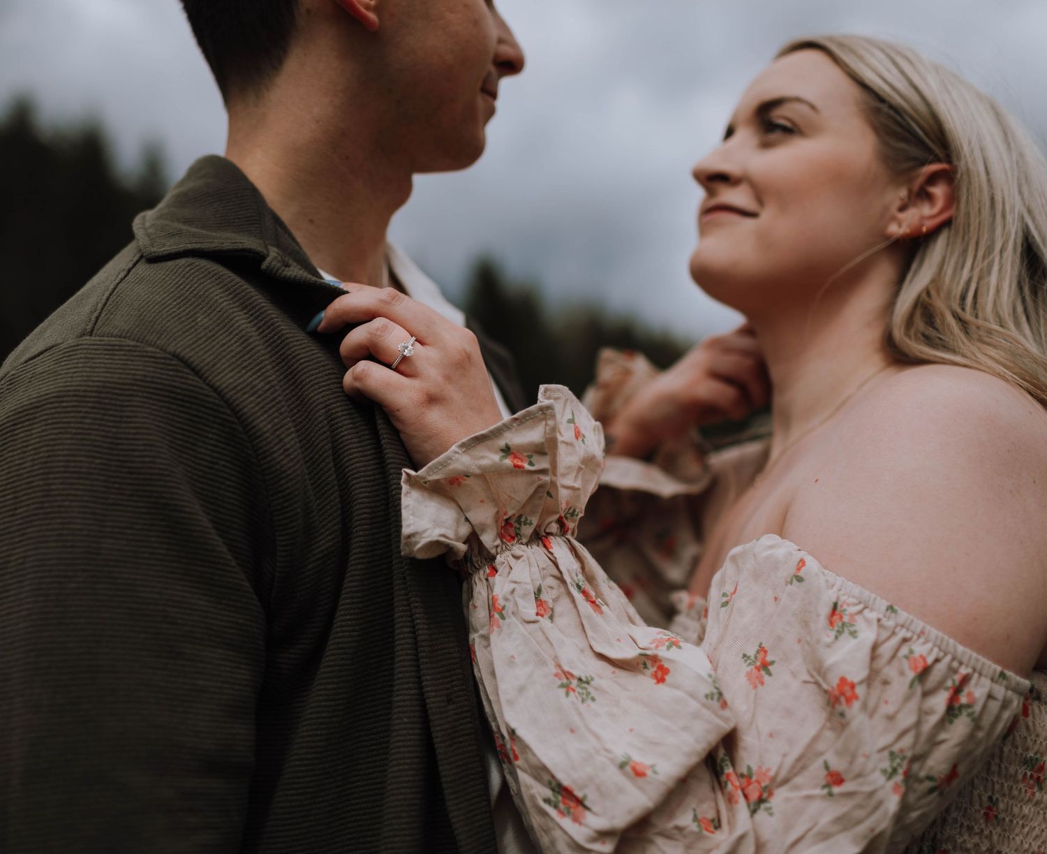 Couple in romantic embrace against forest backdrop with bride wearing floral lace dress and groom in dark suit.
