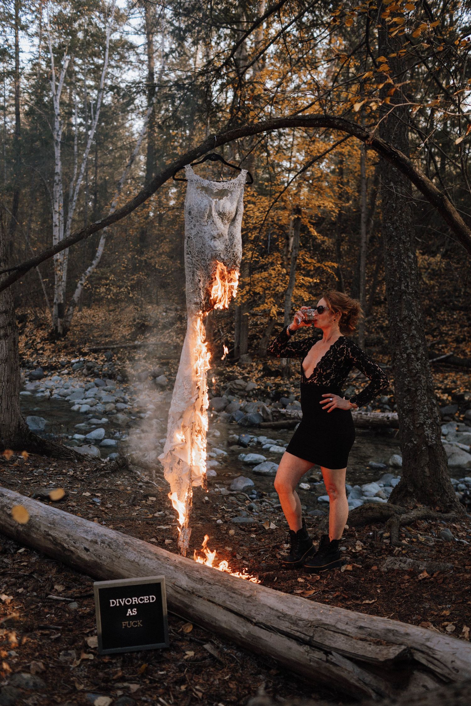 A person in black clothing performs a fire trick with a burning log in an autumn forest setting.