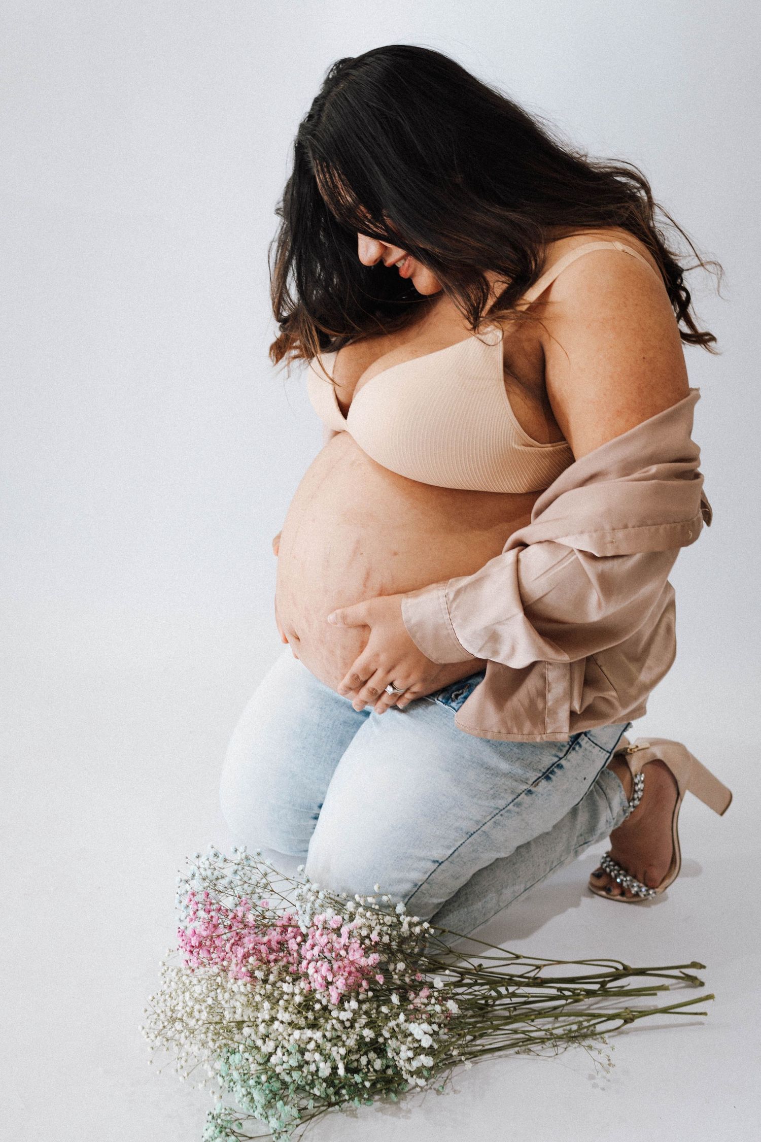 A pregnant woman in light denim and beige cardigan kneels beside colorful dried baby's breath flowers against white background.