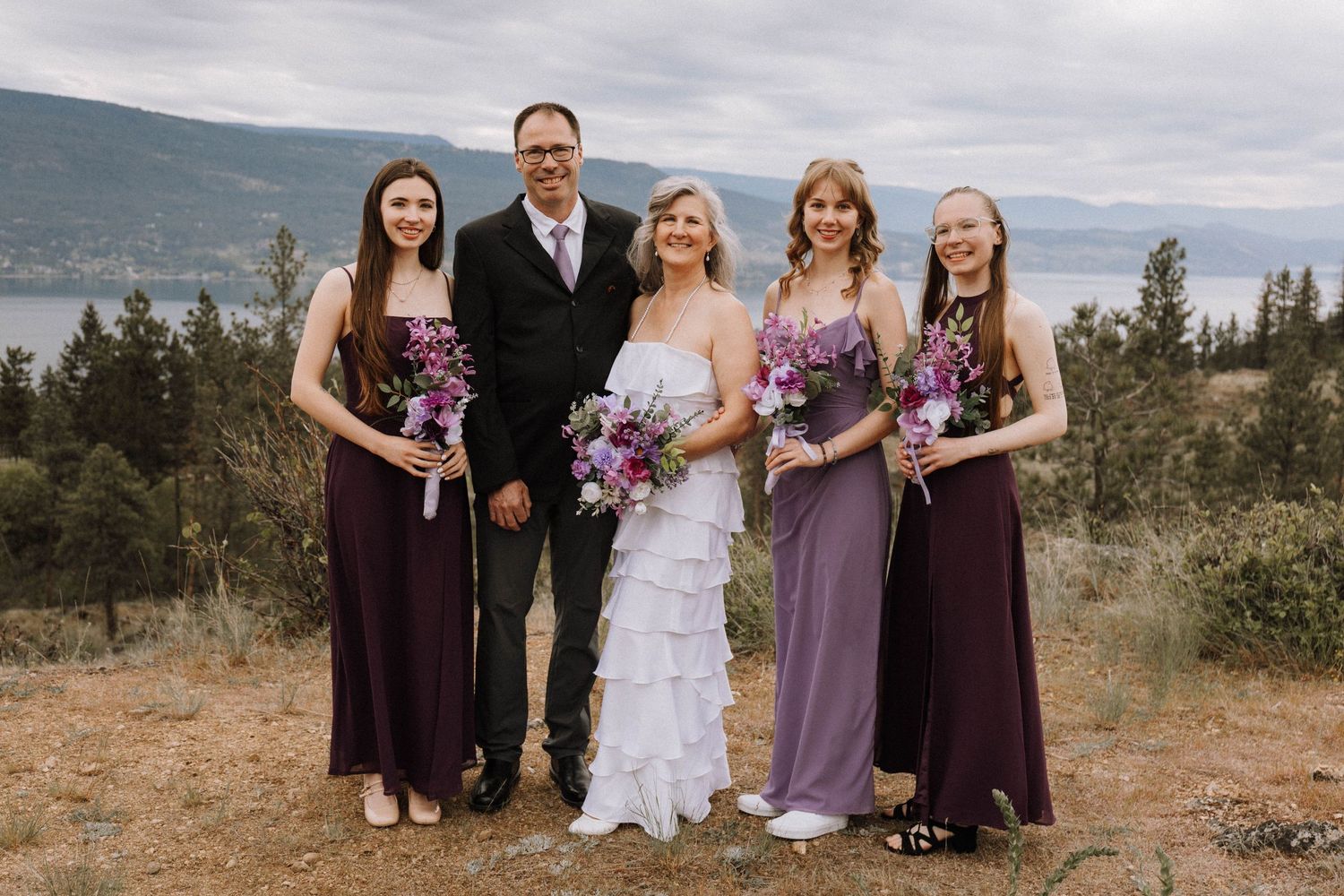 Wedding party poses together on mountain overlook with lake and cloudy skies in background.