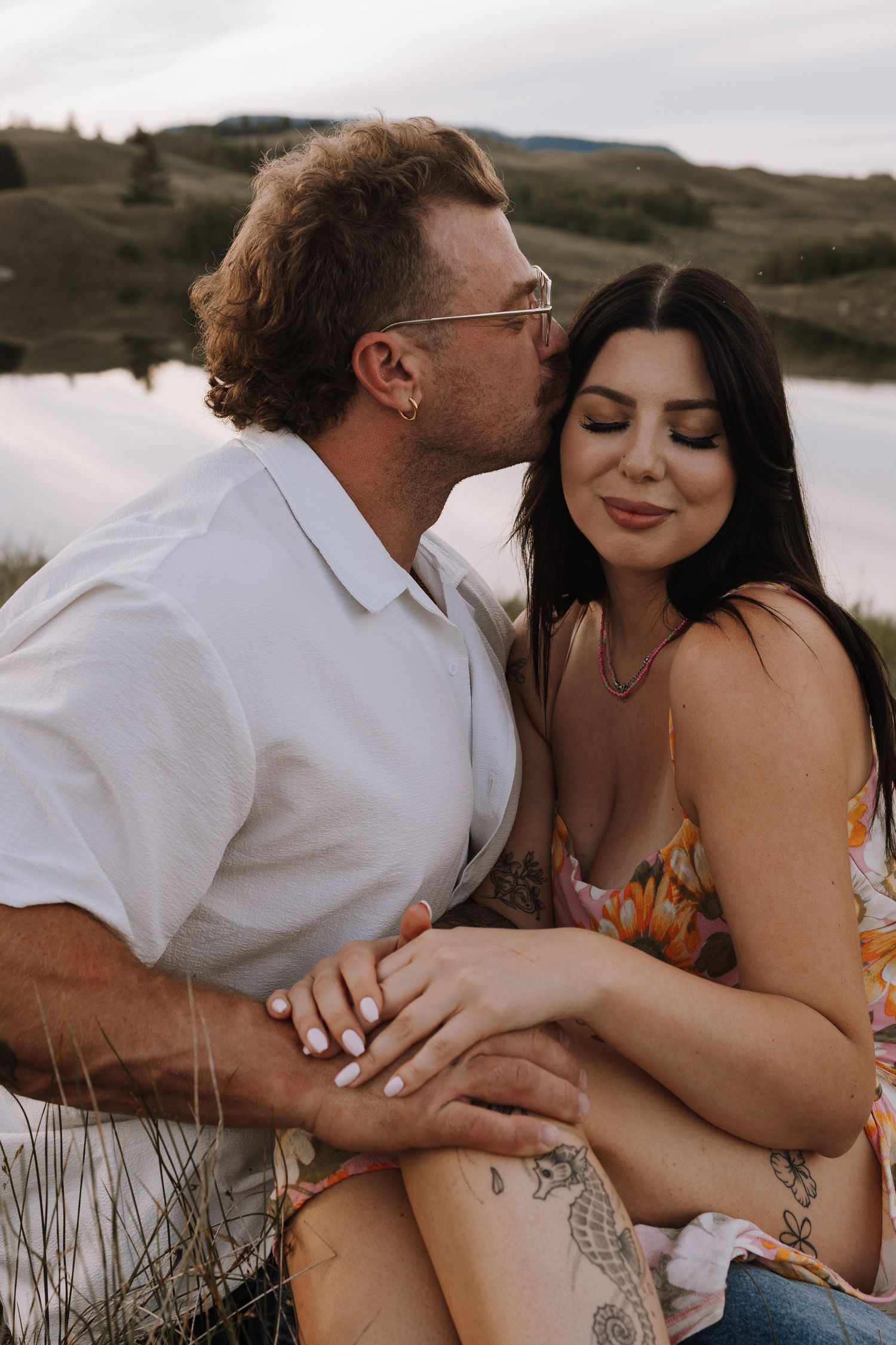 Couple sharing a tender kiss by a scenic lake, woman in floral dress with tattoos, man in white shirt.