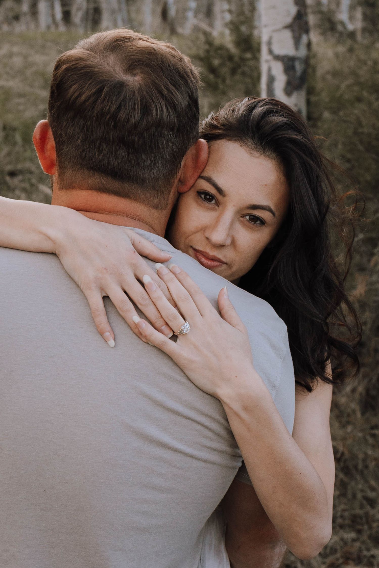 A woman with dark hair hugs a man outdoors, showing an engagement ring, with trees in the background.