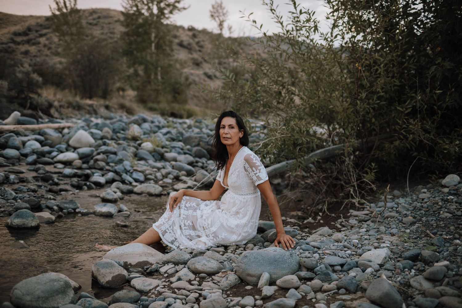 A tranquil scene of someone sitting on rocky terrain near a river at dusk.