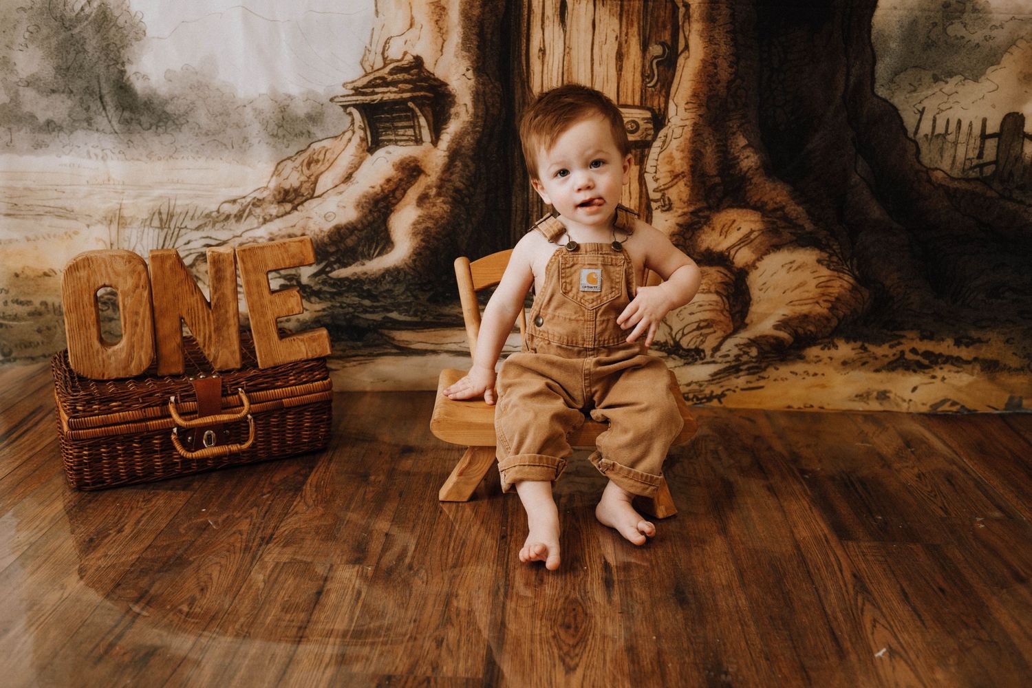 Baby in brown overalls sitting by rustic wooden display for first birthday.