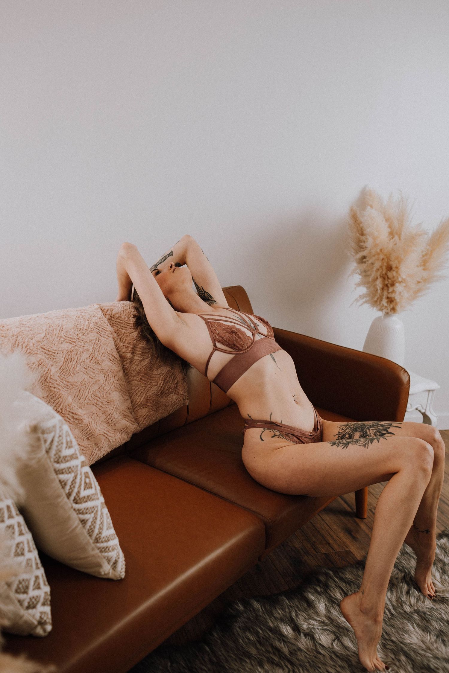 Artistic boudoir shot of a person reclining on a brown leather couch with decorative pillows and pampas grass decor.