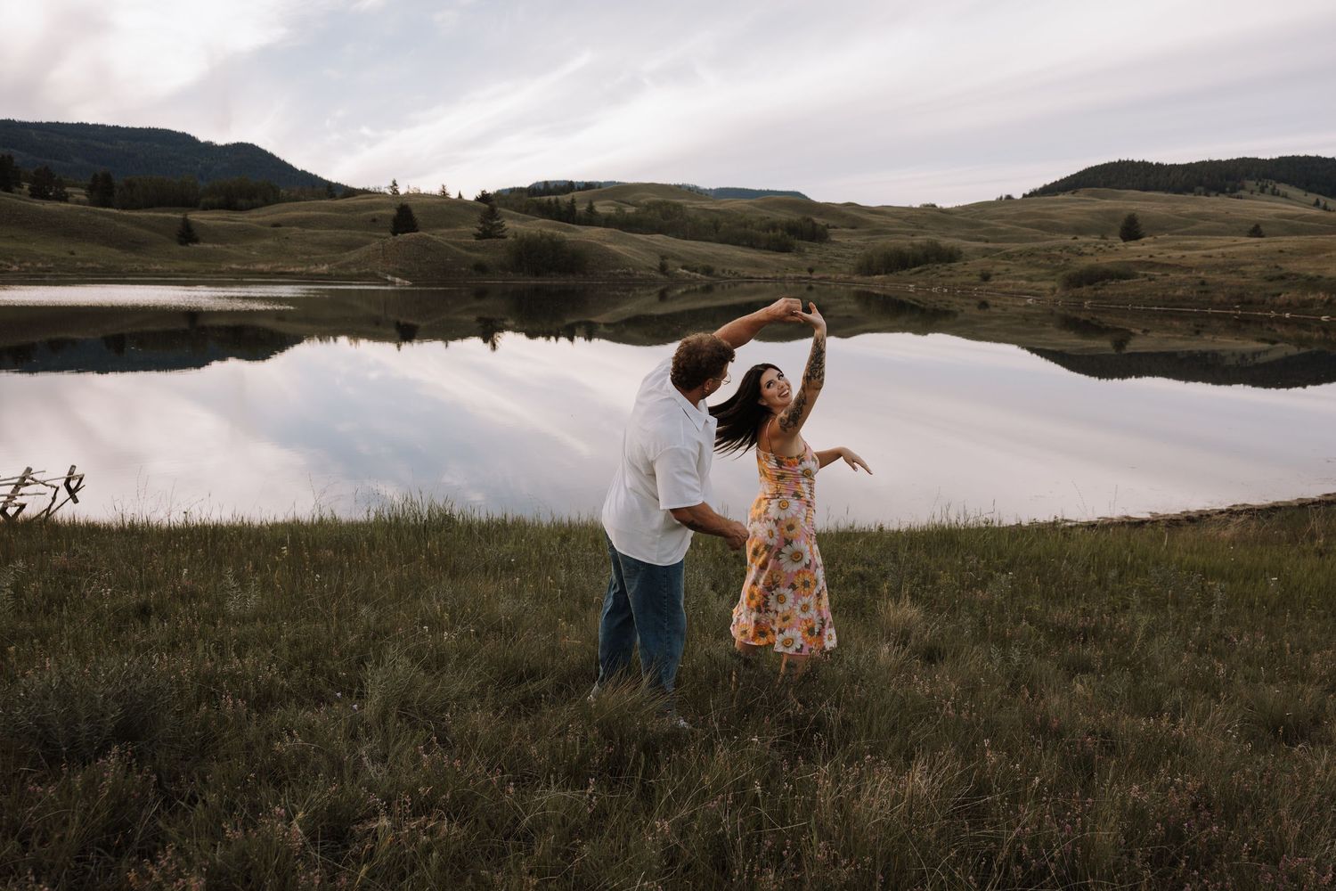 Couple dancing together in a meadow by a reflective mountain lake at dusk, woman in floral dress being twirled.