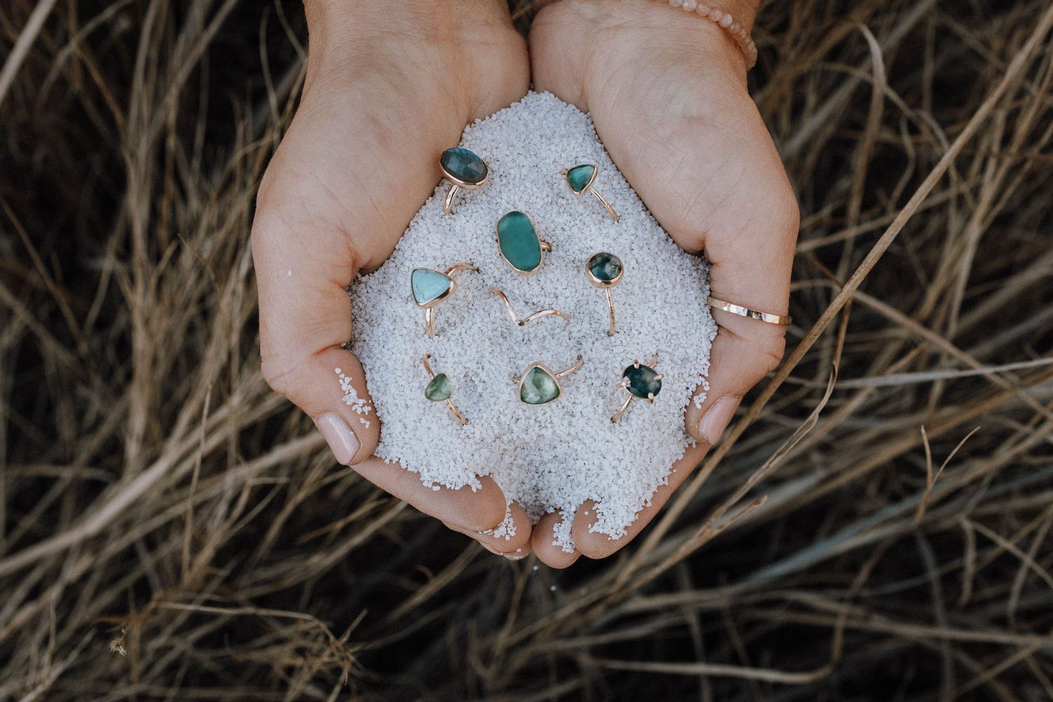 Hands holding white sea salt crystals and small pieces of sea glass against a natural beach grass background.