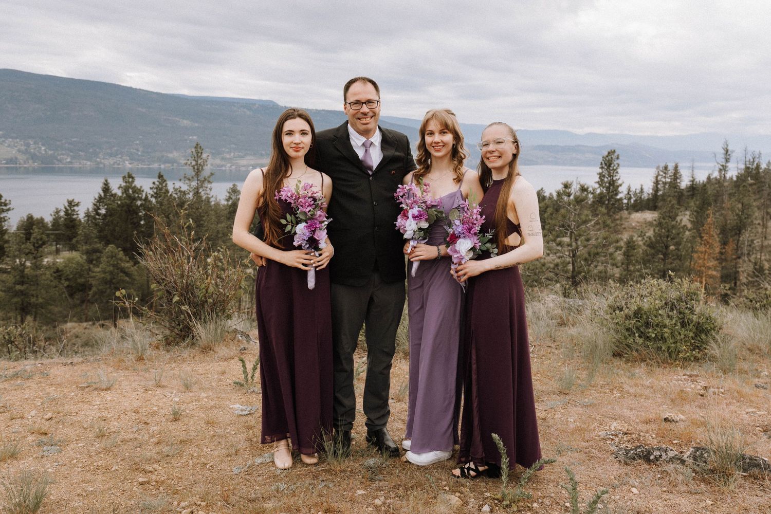 Wedding party wearing purple dresses and black suit pose together on scenic mountainside overlooking lake on cloudy day.