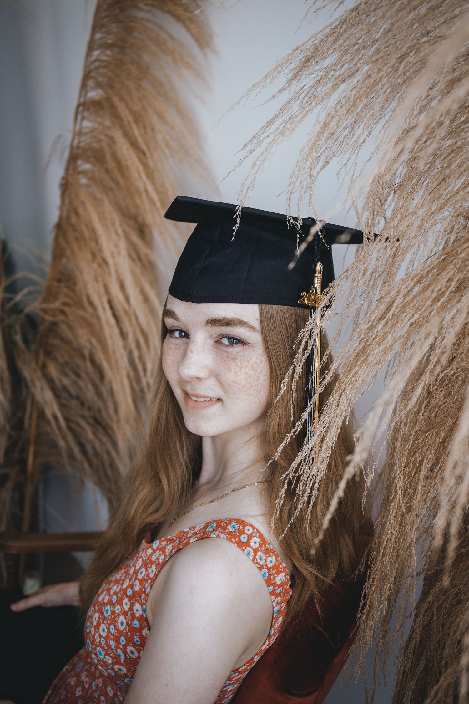 Graduate wearing mortarboard cap and floral dress poses against pampas grass background for graduation photos.