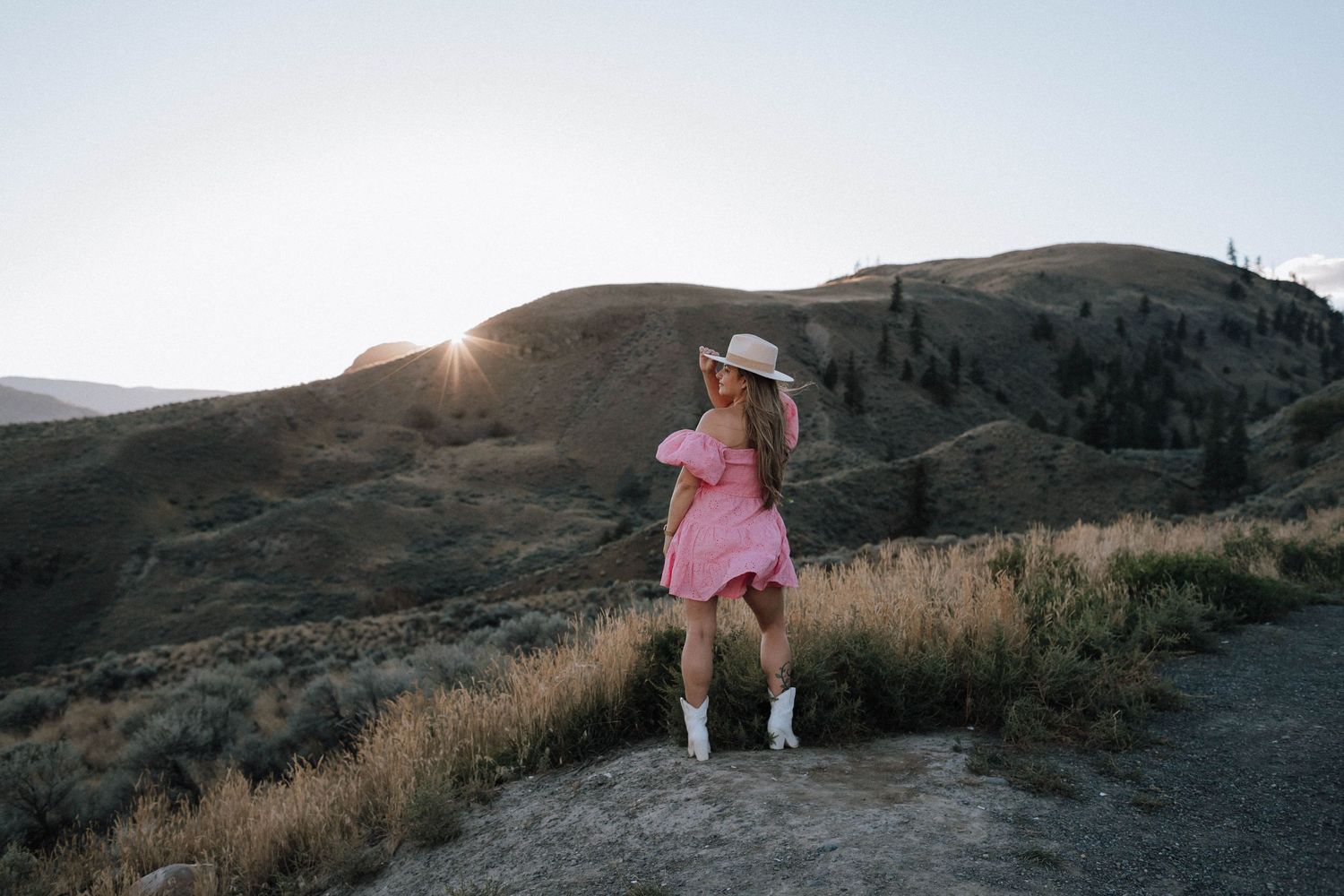 A silhouette poses on a mountain trail at sunset wearing a pink dress and boots.