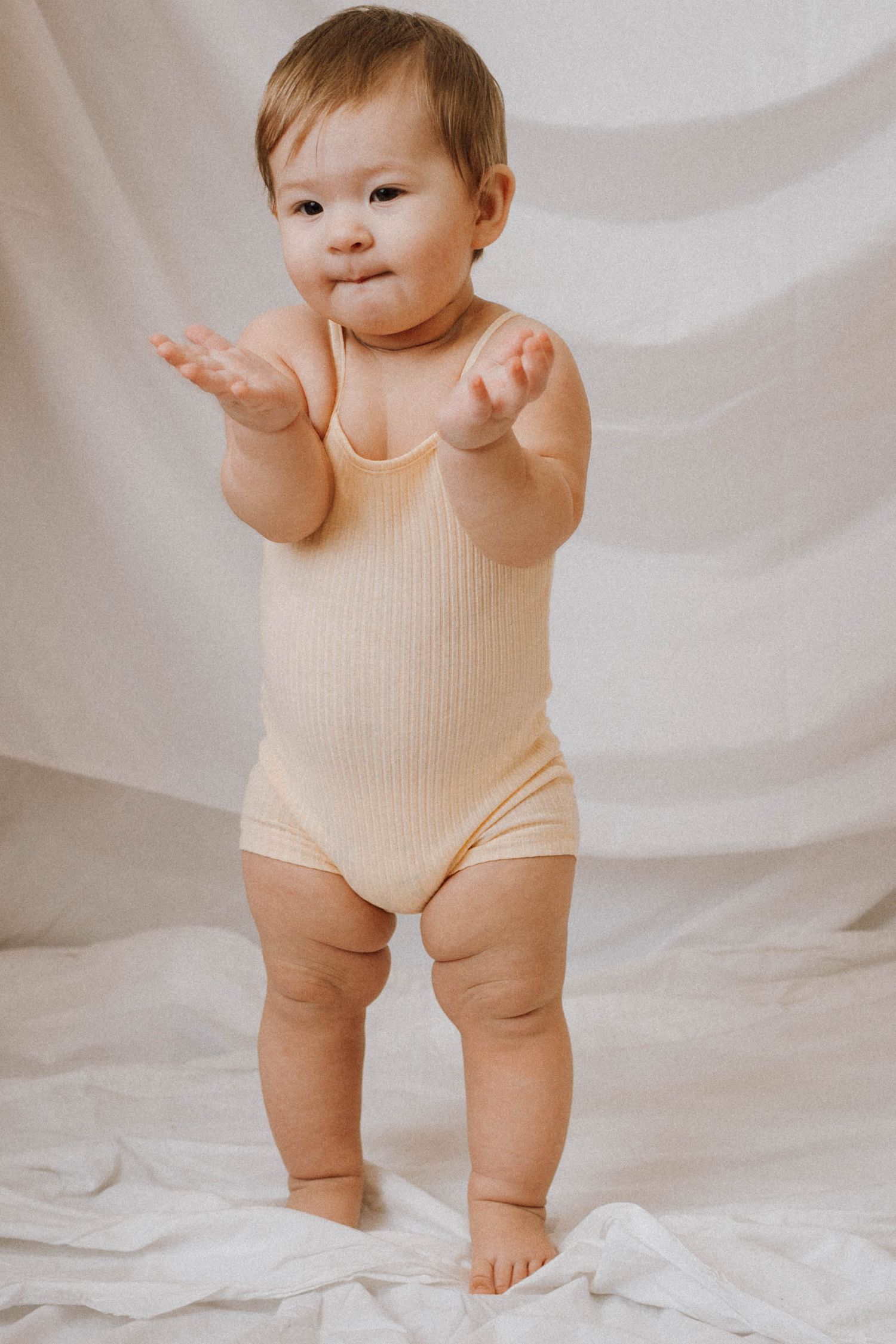 Baby in beige ribbed bodysuit standing on white backdrop with arms raised playfully.