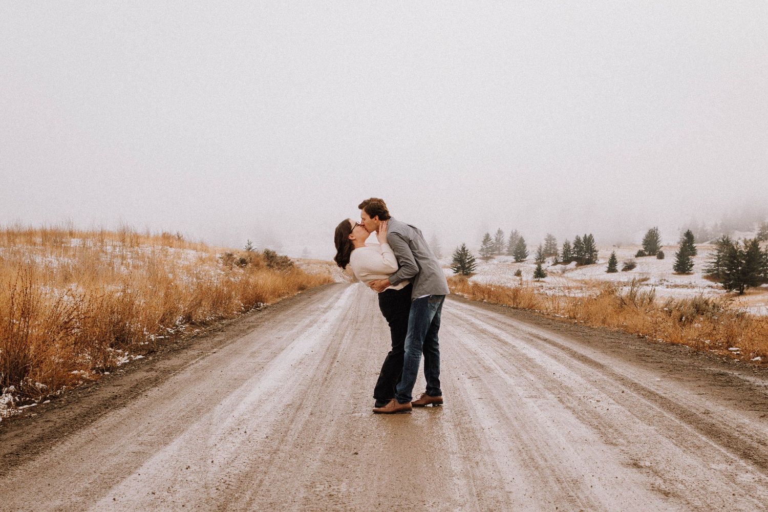 A couple shares a romantic moment on a dirt road surrounded by golden fields and evergreen trees in the distance.
