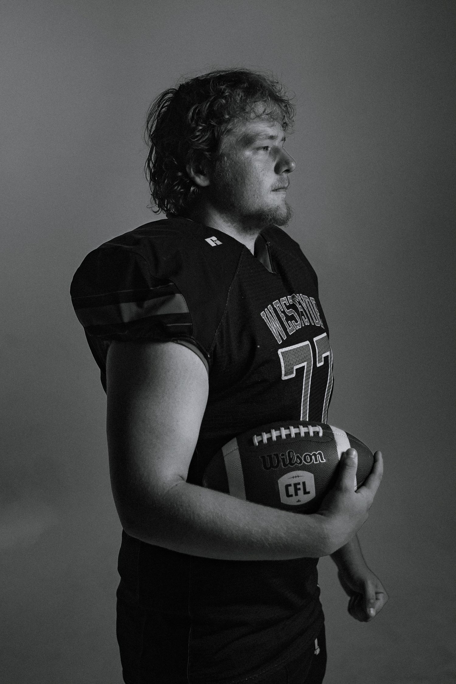 Black and white photo of a football player in jersey number 77 holding a CFL Wilson football.