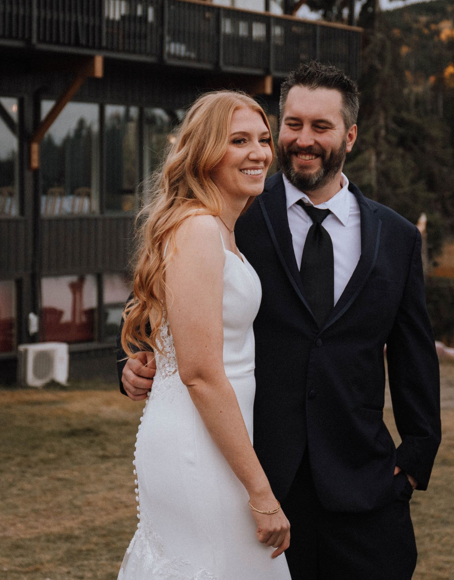 A wedding couple embraces outdoors with the bride in a fitted white gown and groom in dark suit.