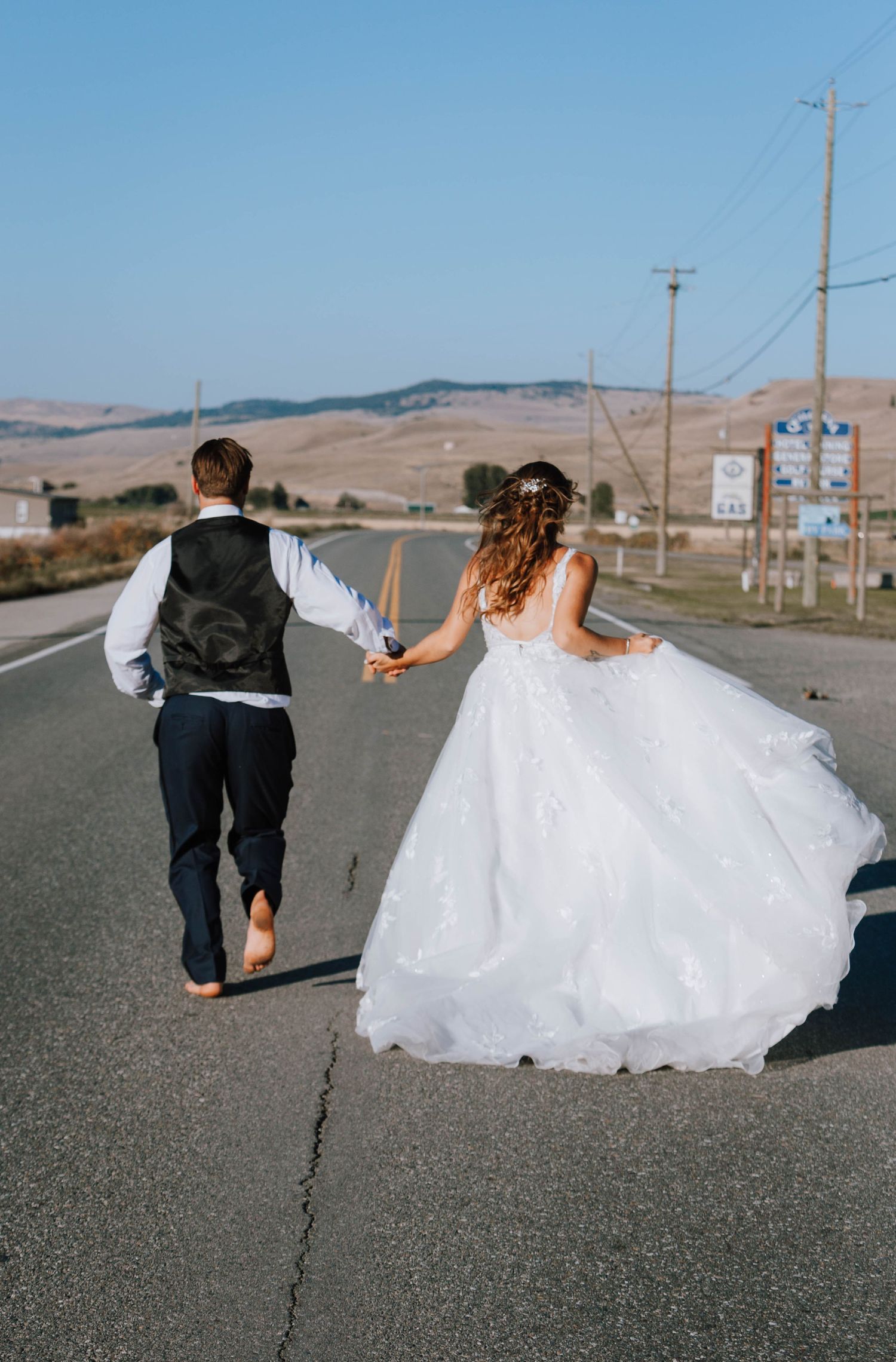 Newlyweds walk barefoot down an empty road holding hands against mountain backdrop.