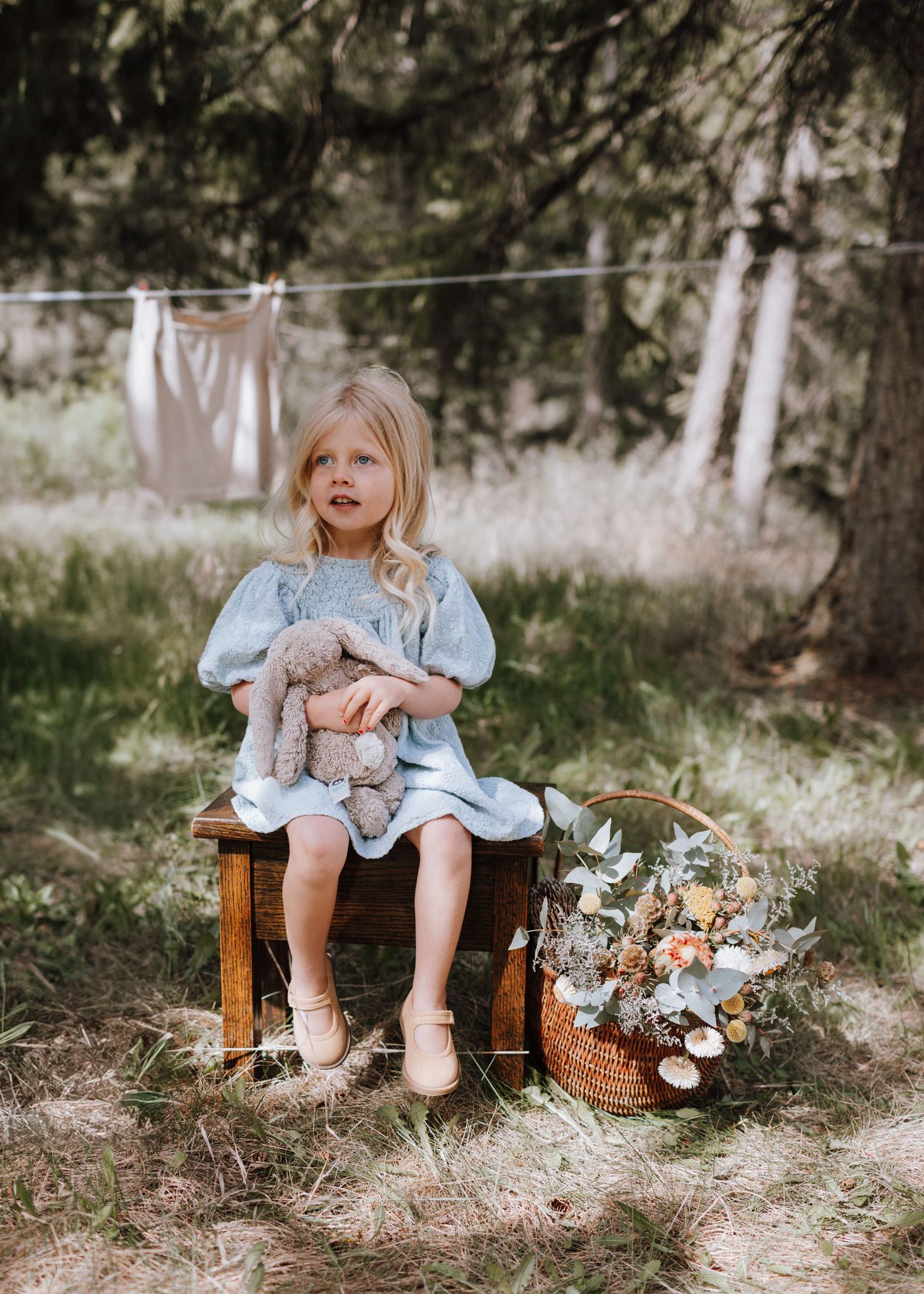 A child in a blue dress sits on a wooden bench surrounded by dried flowers and natural outdoor scenery.
