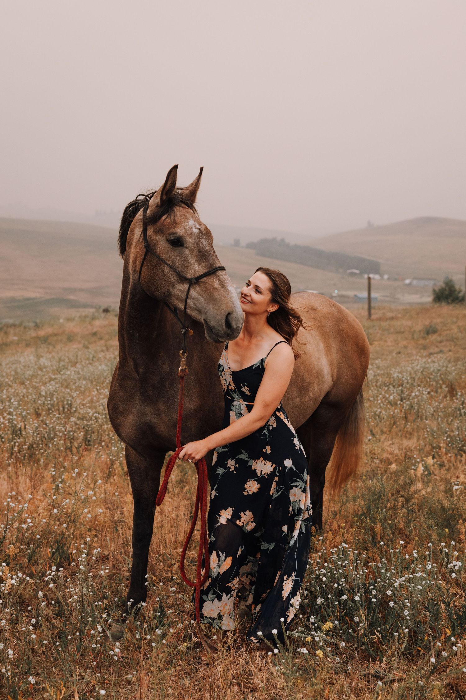Woman in floral dress smiling beside a brown horse in a scenic field with misty hills in the background.