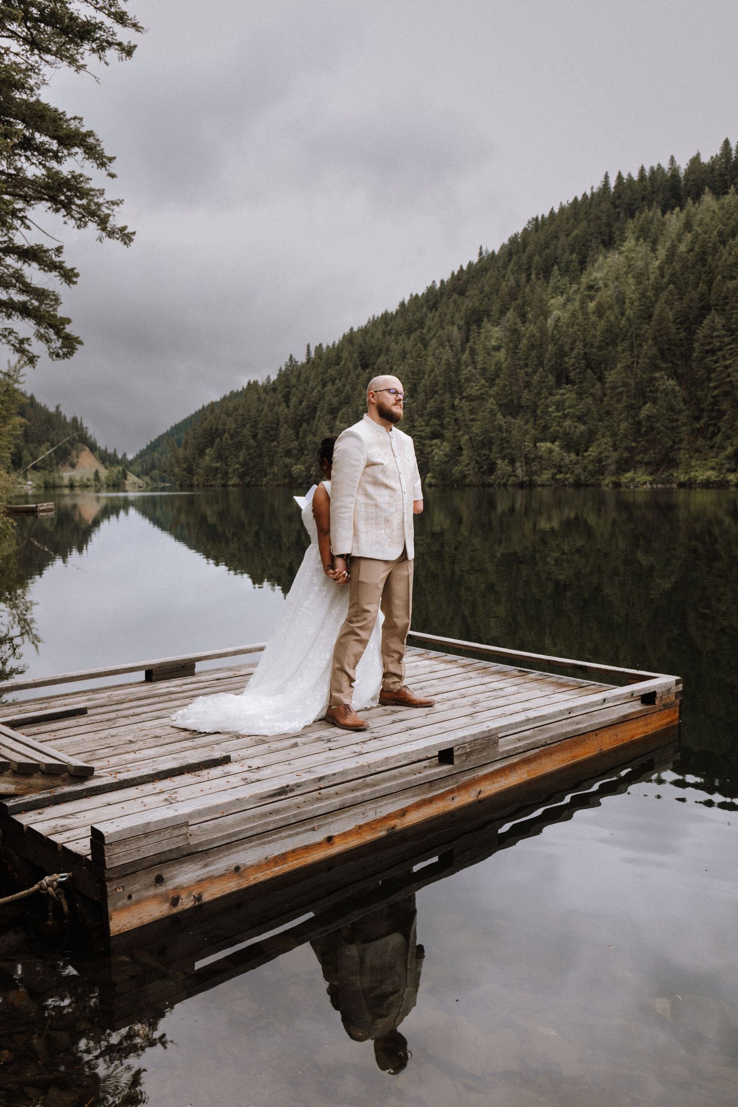 Someone in light-colored clothing stands on a wooden dock by a serene lake surrounded by evergreen-covered hills on a cloudy day.