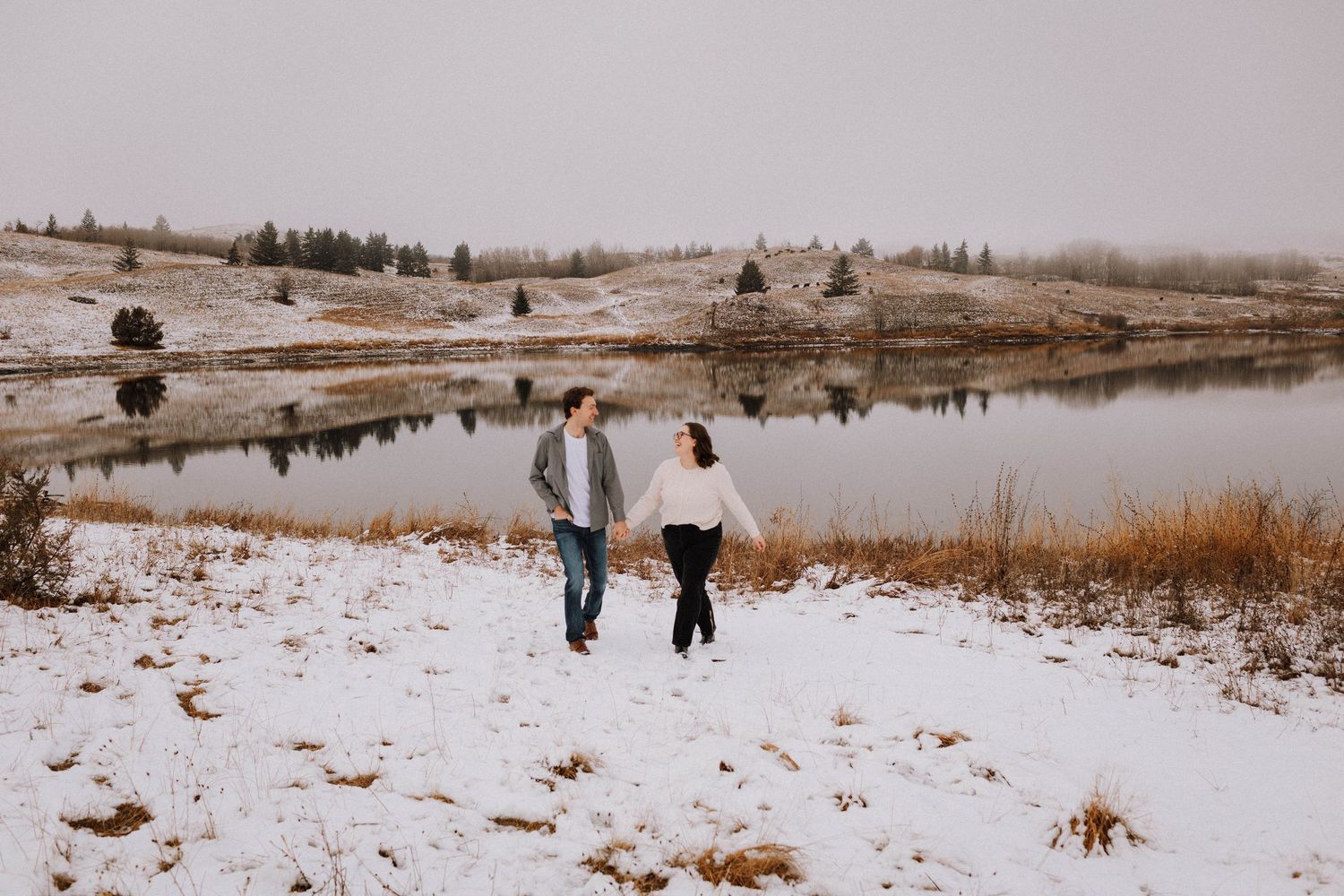 People walk along a snowy path through a winter landscape with brown grass and scattered trees in the distance.